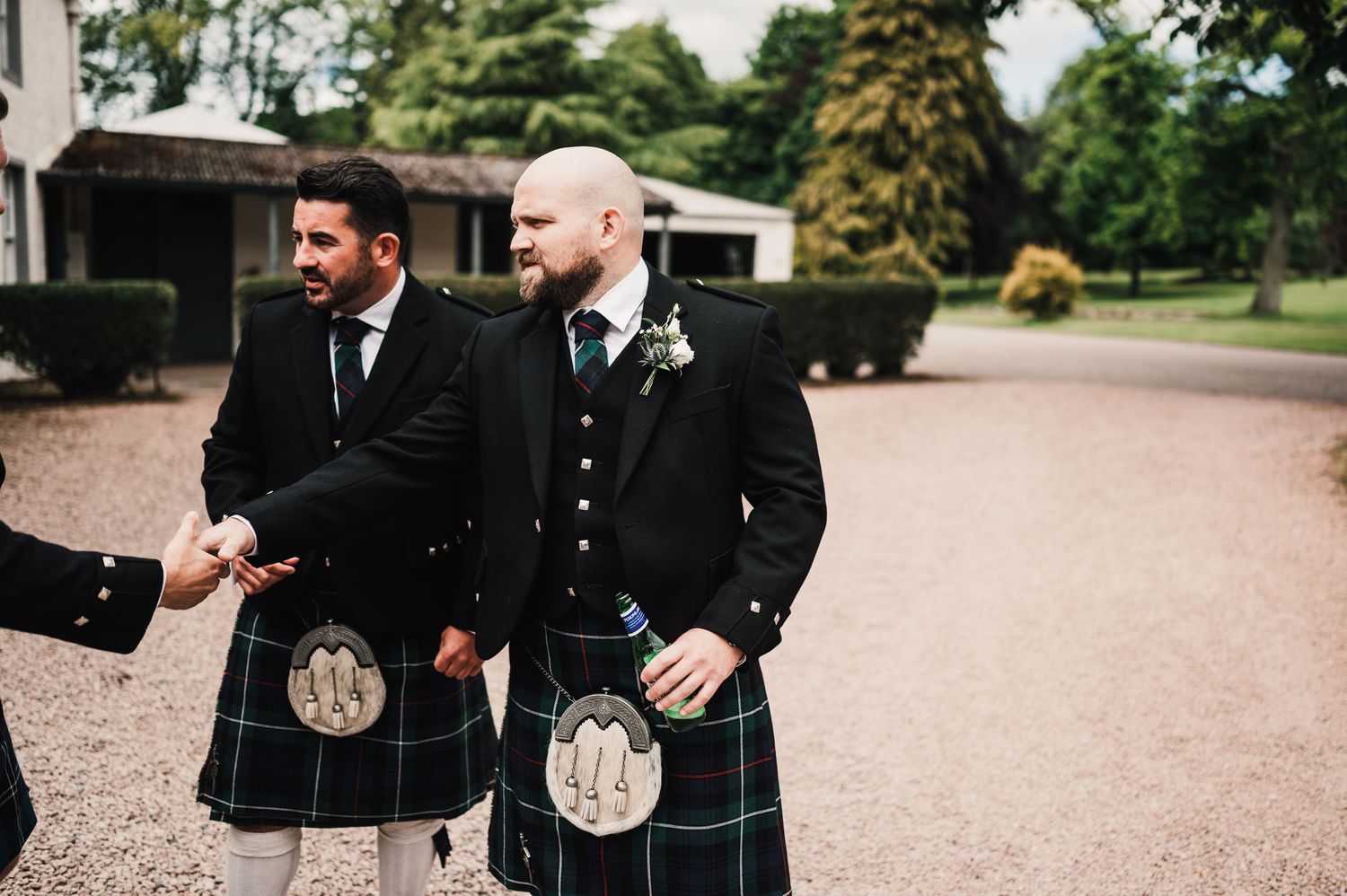 Two men in traditional Scottish kilts and formal jackets stand on a gravel driveway.