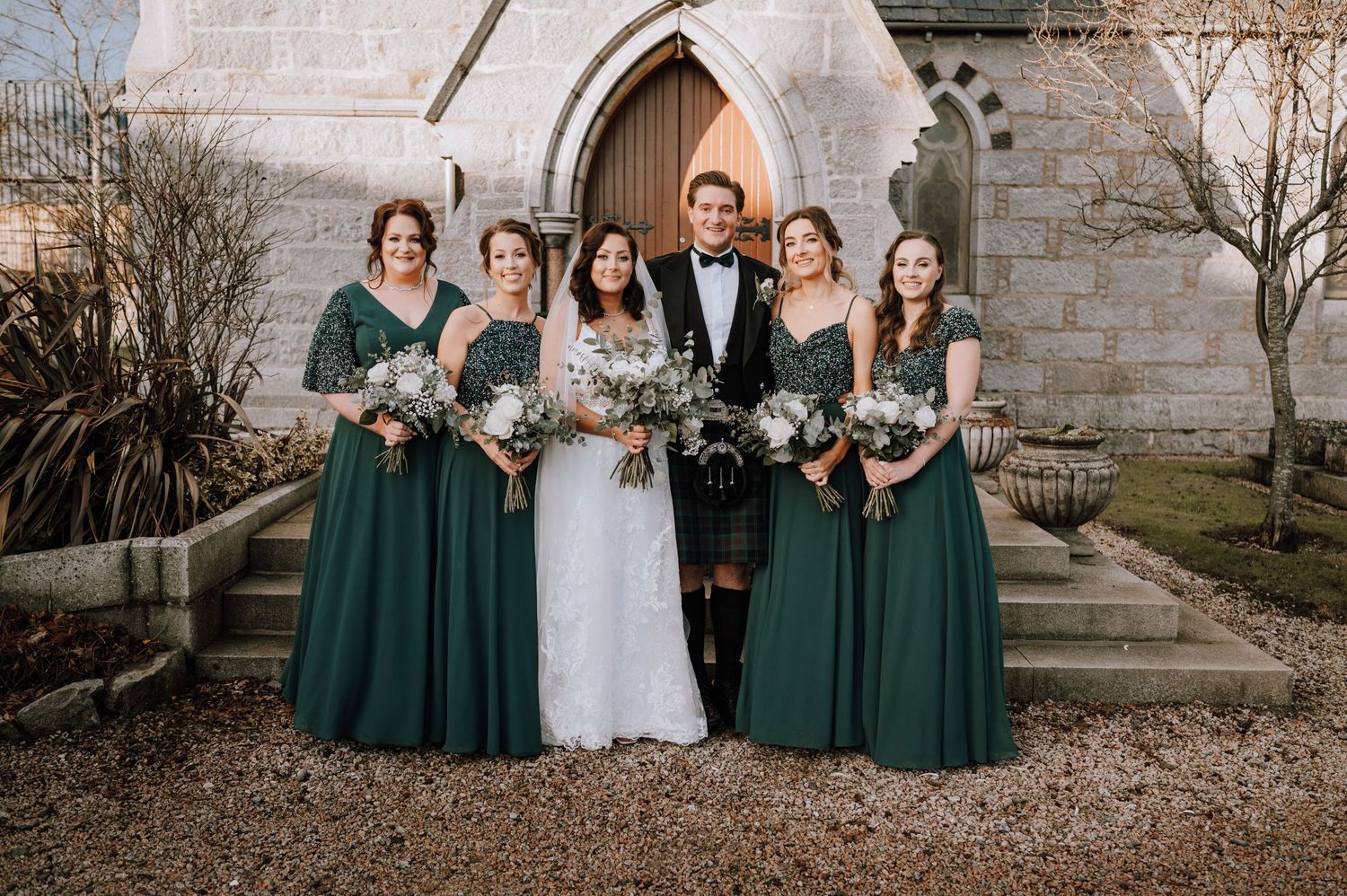 Bridal party wearing elegant emerald green dresses pose together on stone church steps.