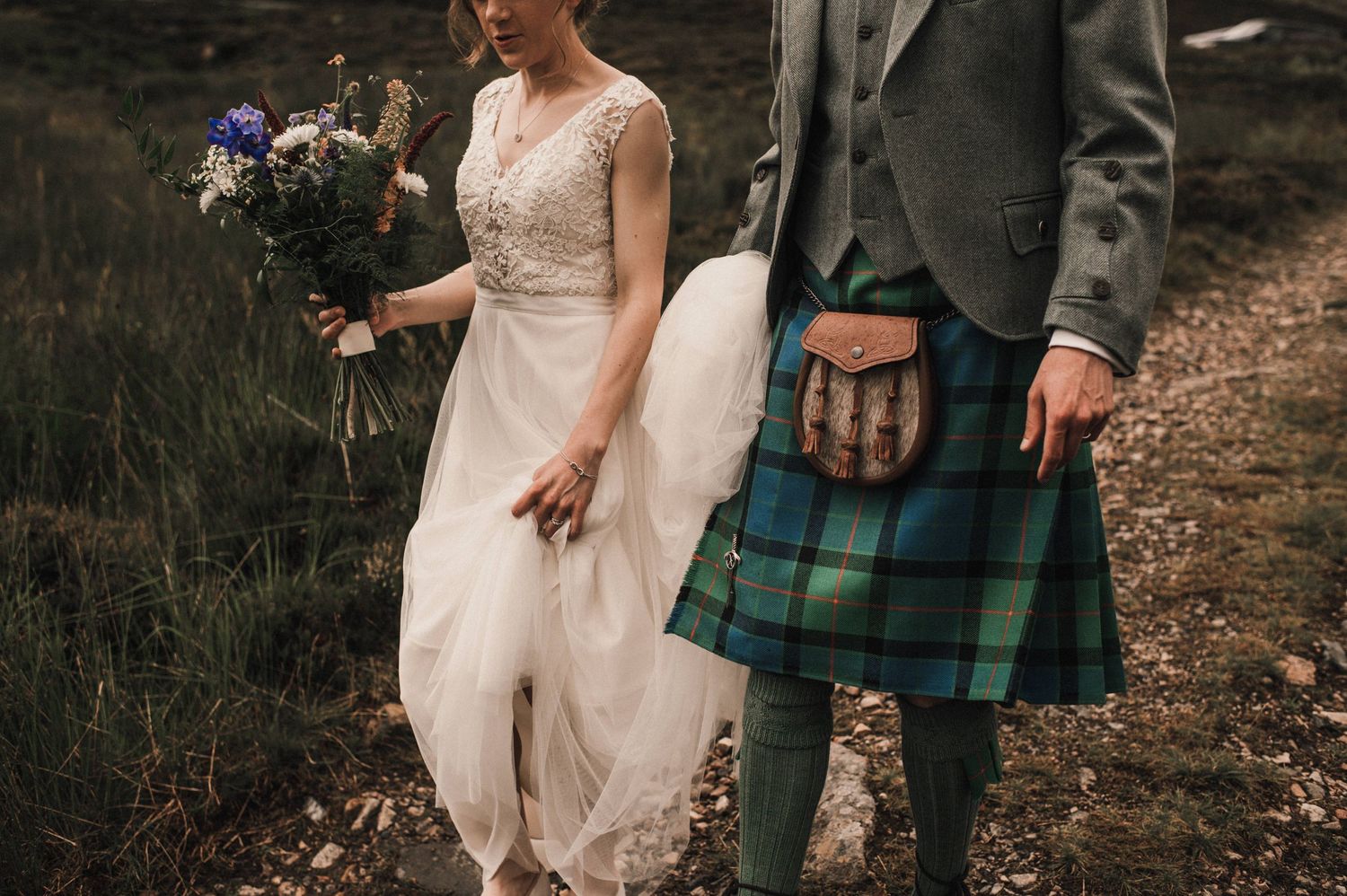 Bride in white dress and groom in kilt stand together on Scottish moorland.