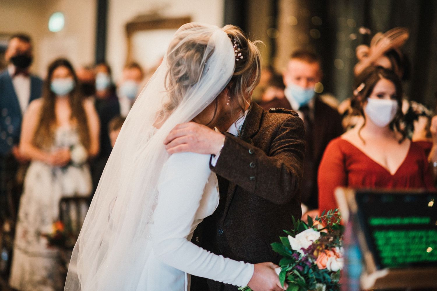 Emotional wedding ceremony moment captured indoors with guests wearing protective masks.