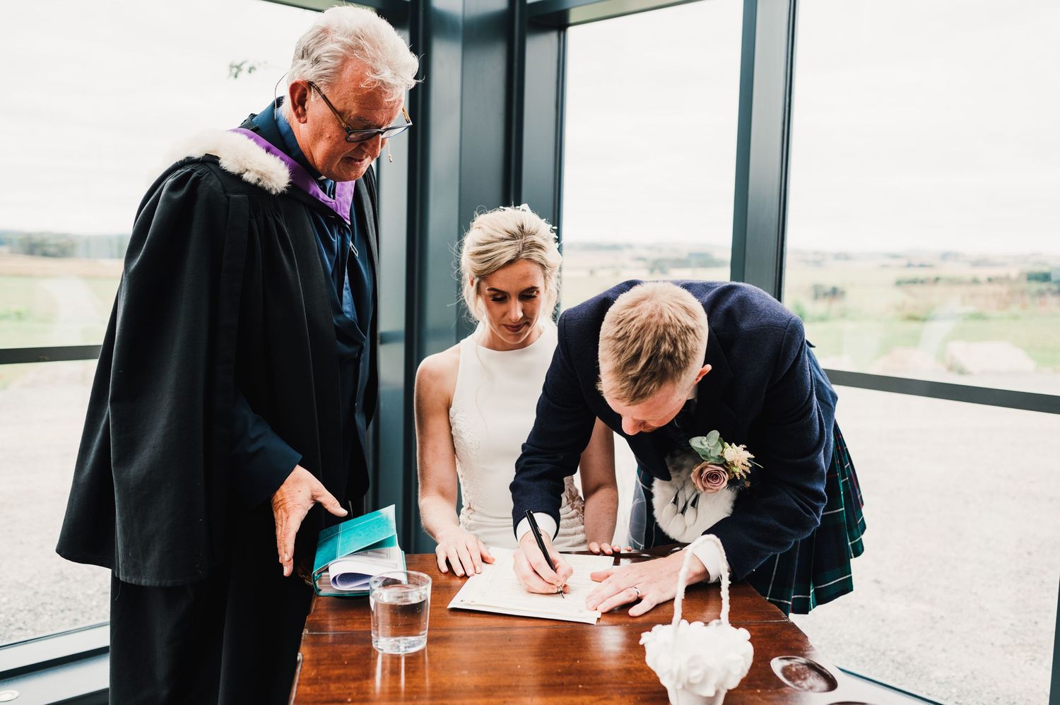 Wedding party signing official documents at a glass-walled venue overlooking countryside.