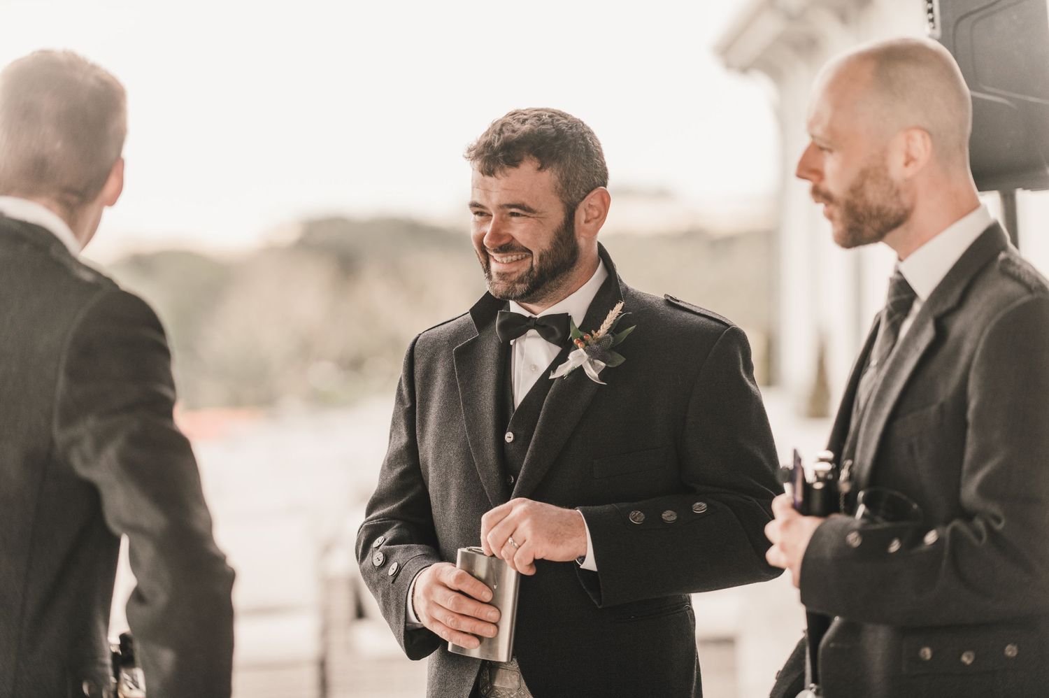 Groomsmen in formal attire sharing drinks and conversation at an outdoor wedding.