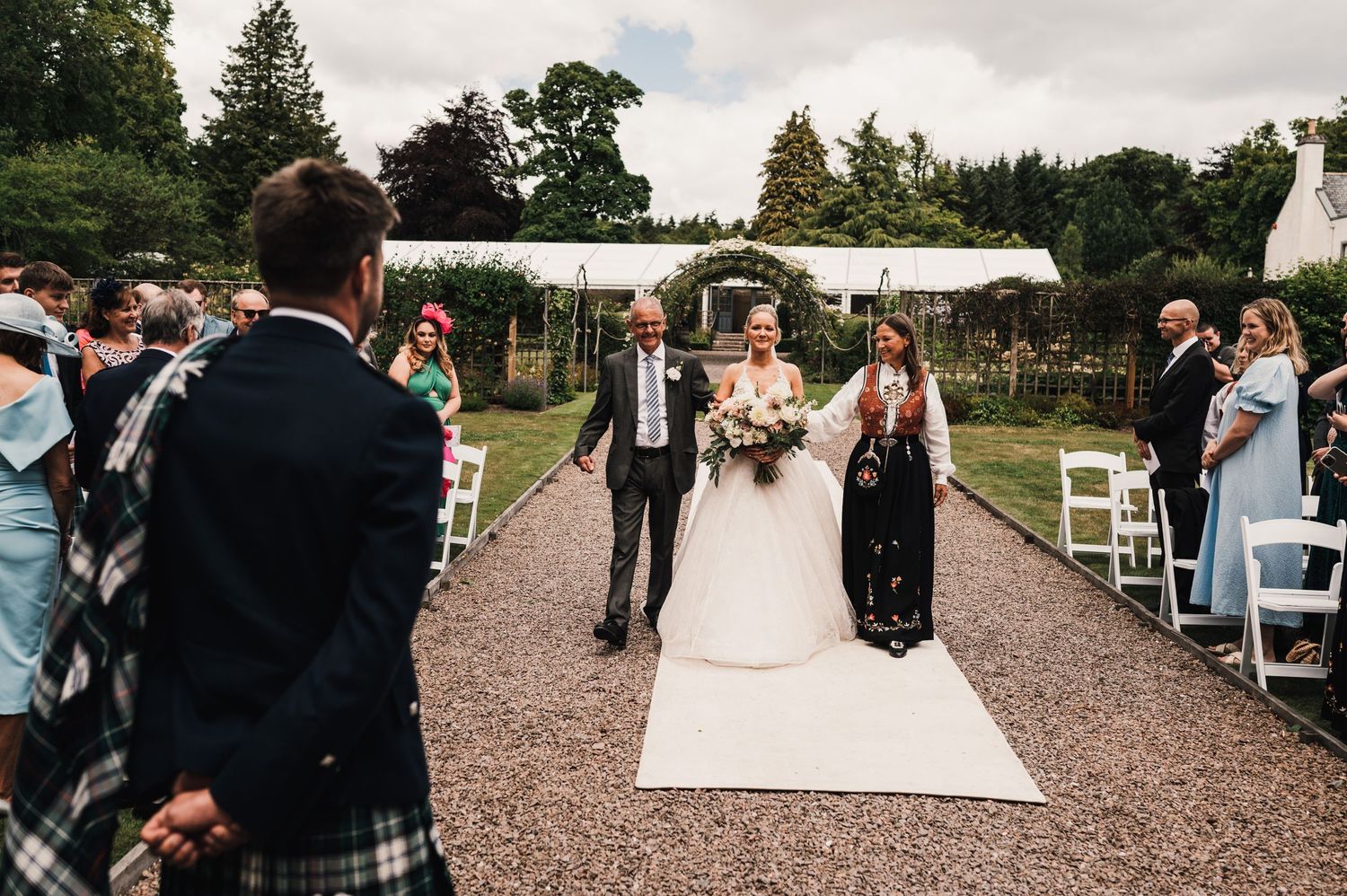 Wedding procession on gravel path with floral arch and seated guests.
