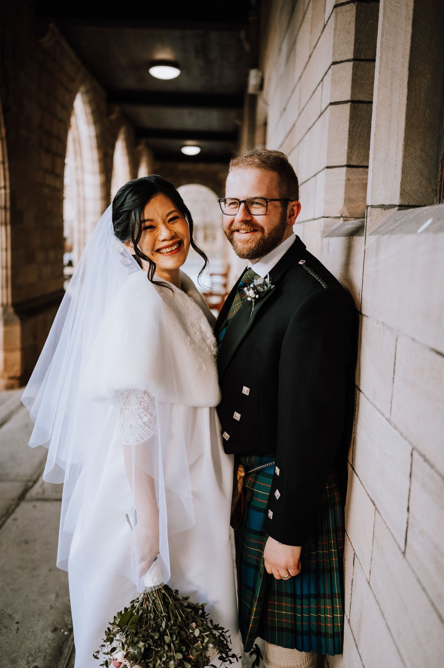 Wedding couple poses in formal attire with Scottish kilt under stone church archway.