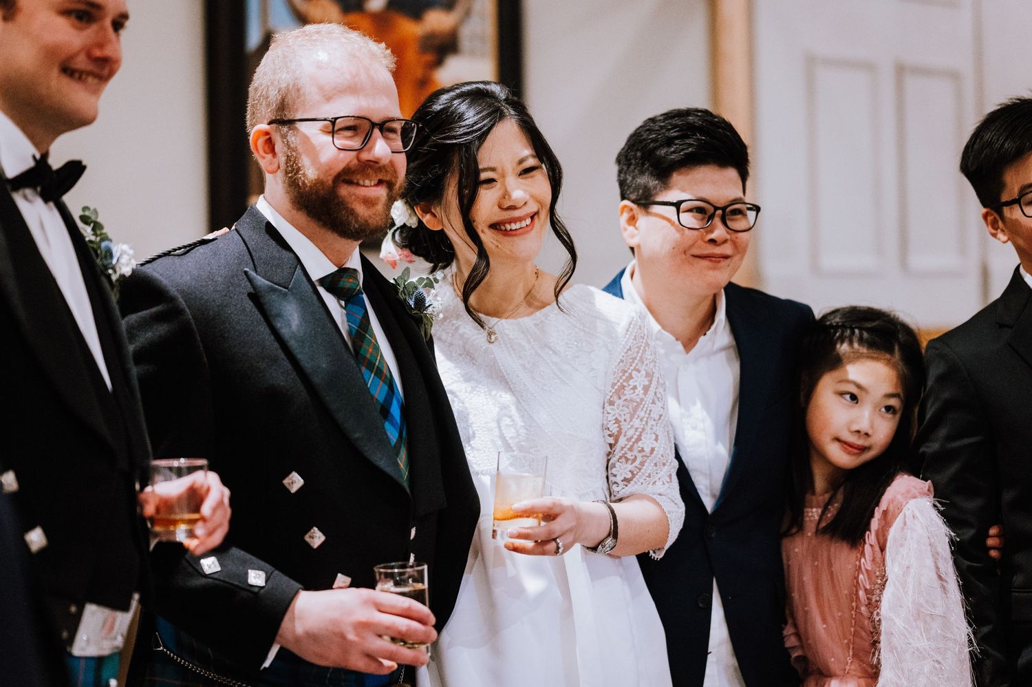 Wedding party members pose together in formal attire including a traditional kilt and white dress.