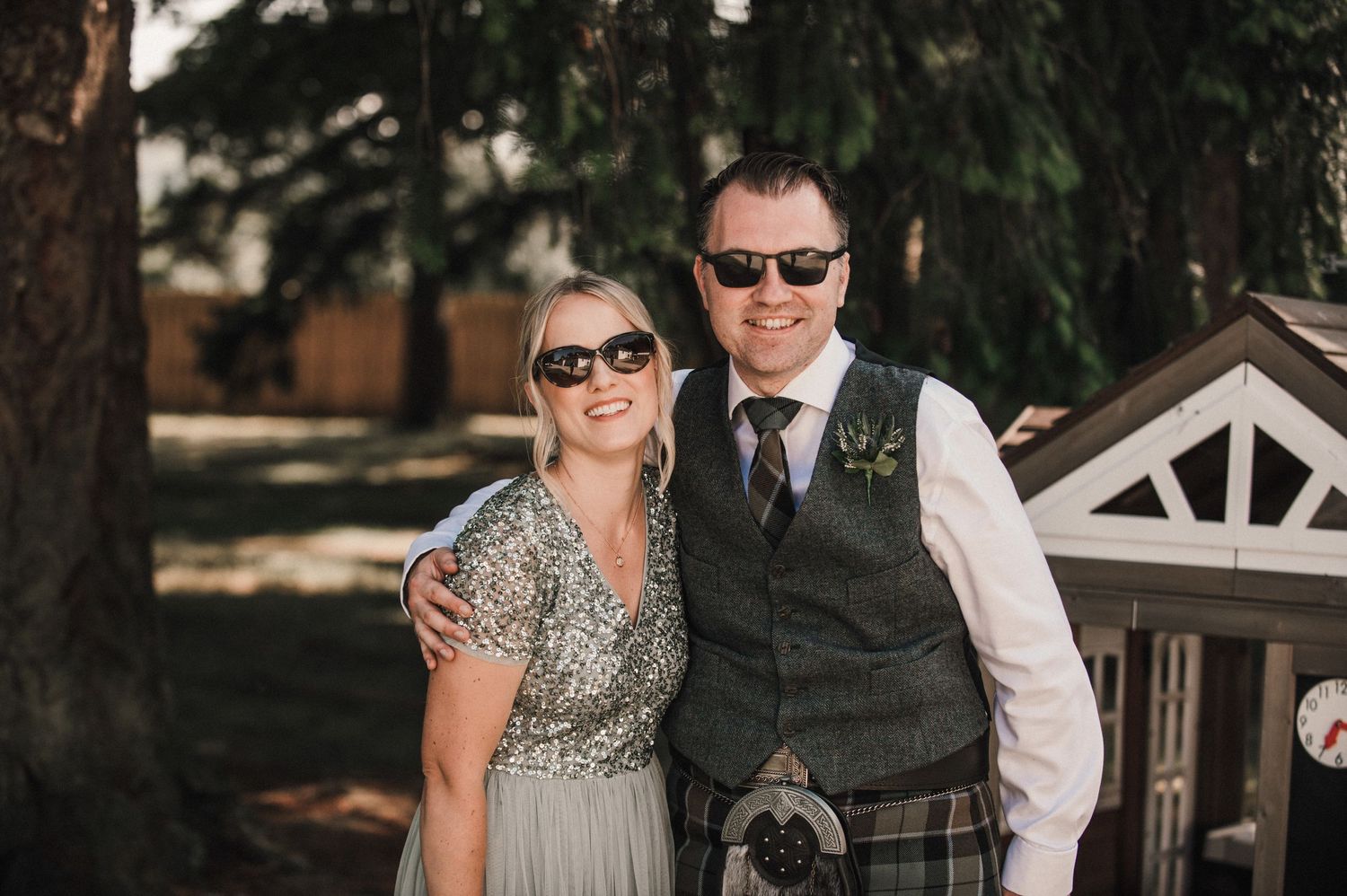 A couple in wedding attire poses outdoors wearing sunglasses with traditional Scottish elements.