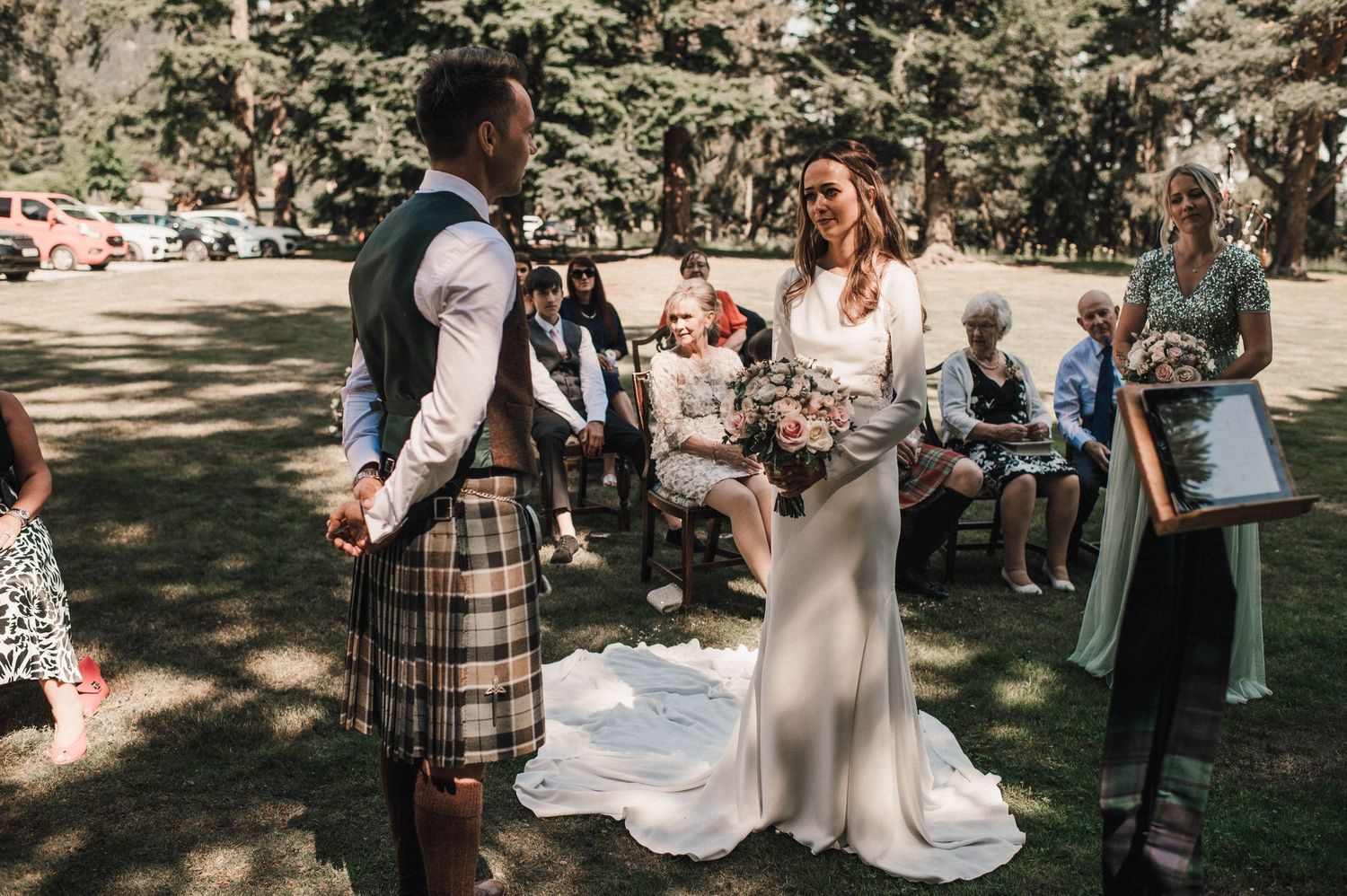 A couple exchanges vows during a wedding ceremony while guests watch in a woodland setting.