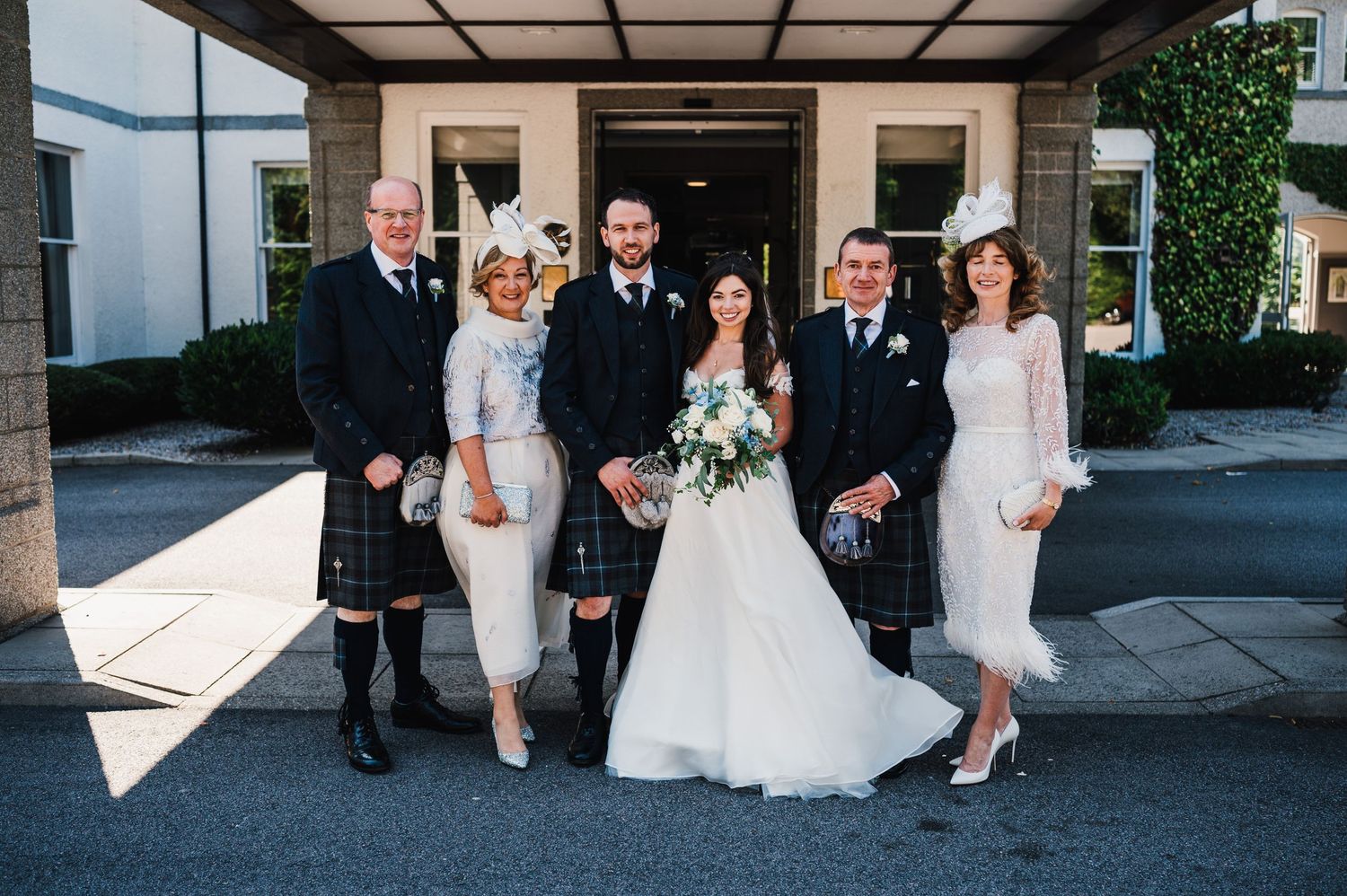 Small family group poses for formal wedding photo outside venue entrance in Scottish formal wear.