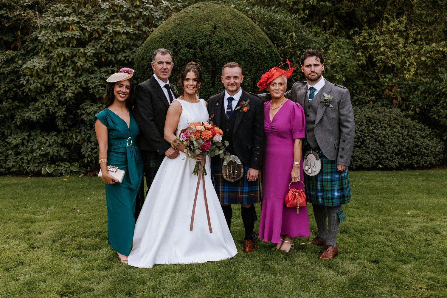 Wedding party members arranged for formal portraits on a manicured lawn with hedges in background.