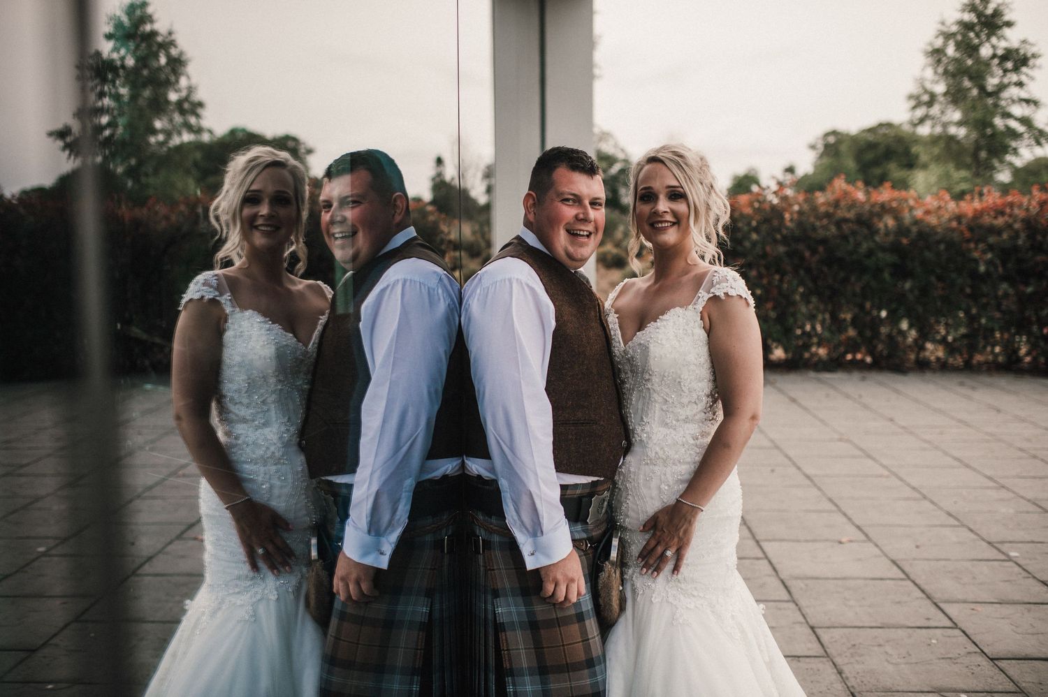 Two couples in wedding attire pose together on outdoor terrace.