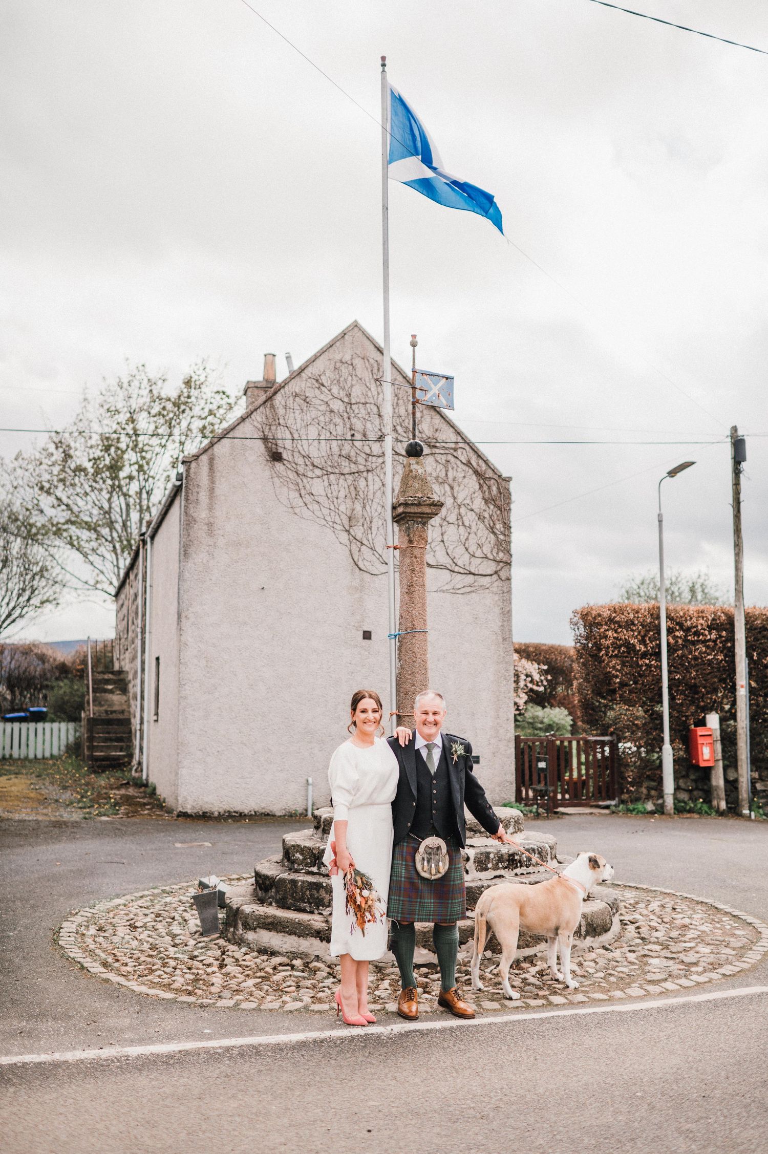 Couple with dog posing at stone monument under Scottish flag.