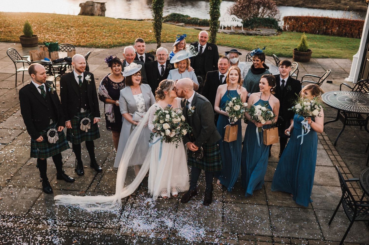 Wedding group photo featuring Scottish kilts and blue bridesmaid dresses on a stone patio.