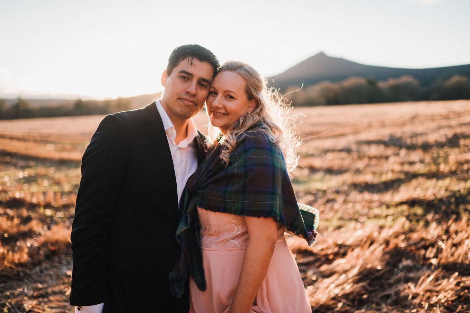 A couple poses together in warm sunlight on a hillside during sunset.