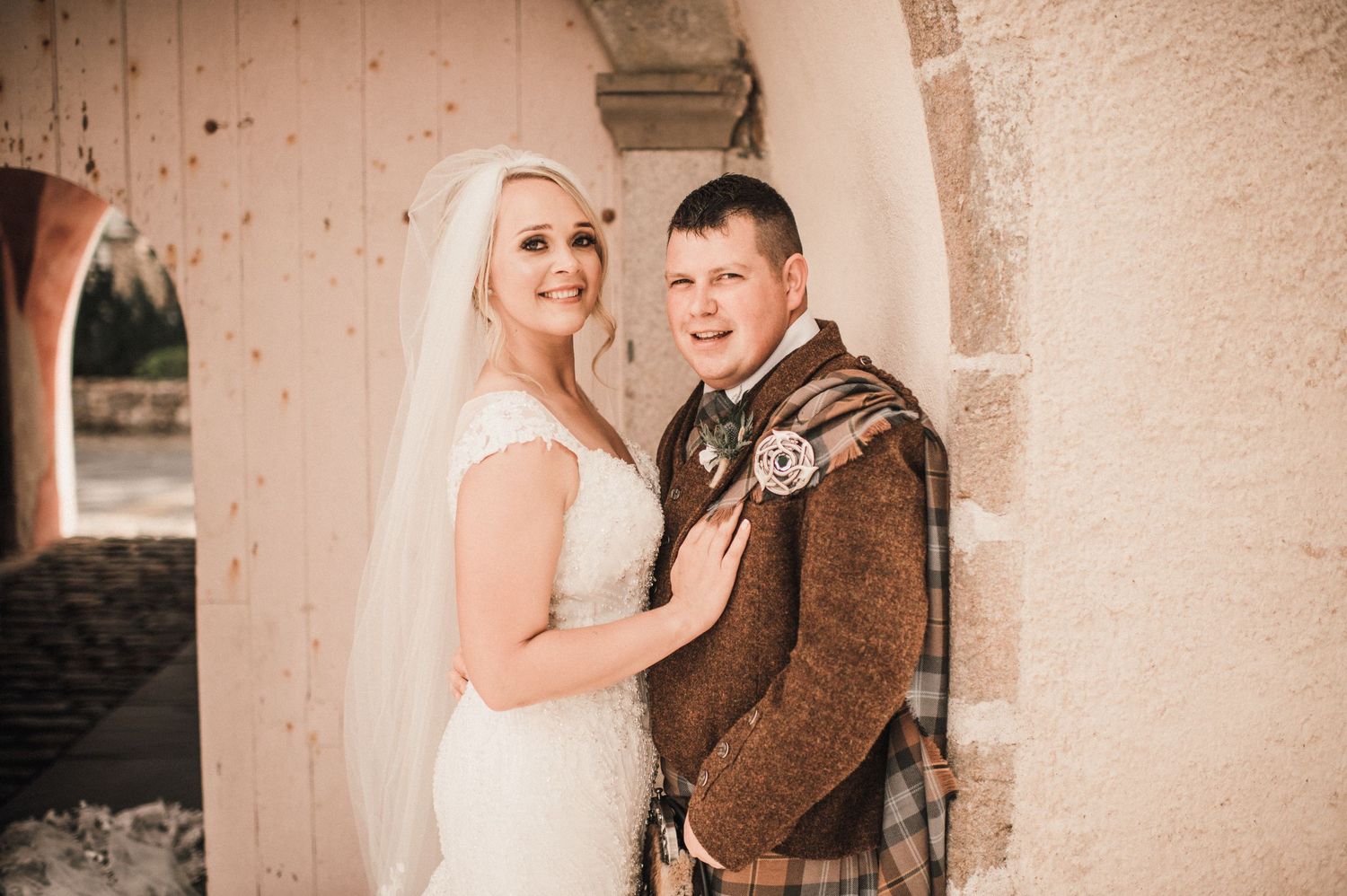A couple in wedding attire poses under an arched stone doorway on their wedding day.