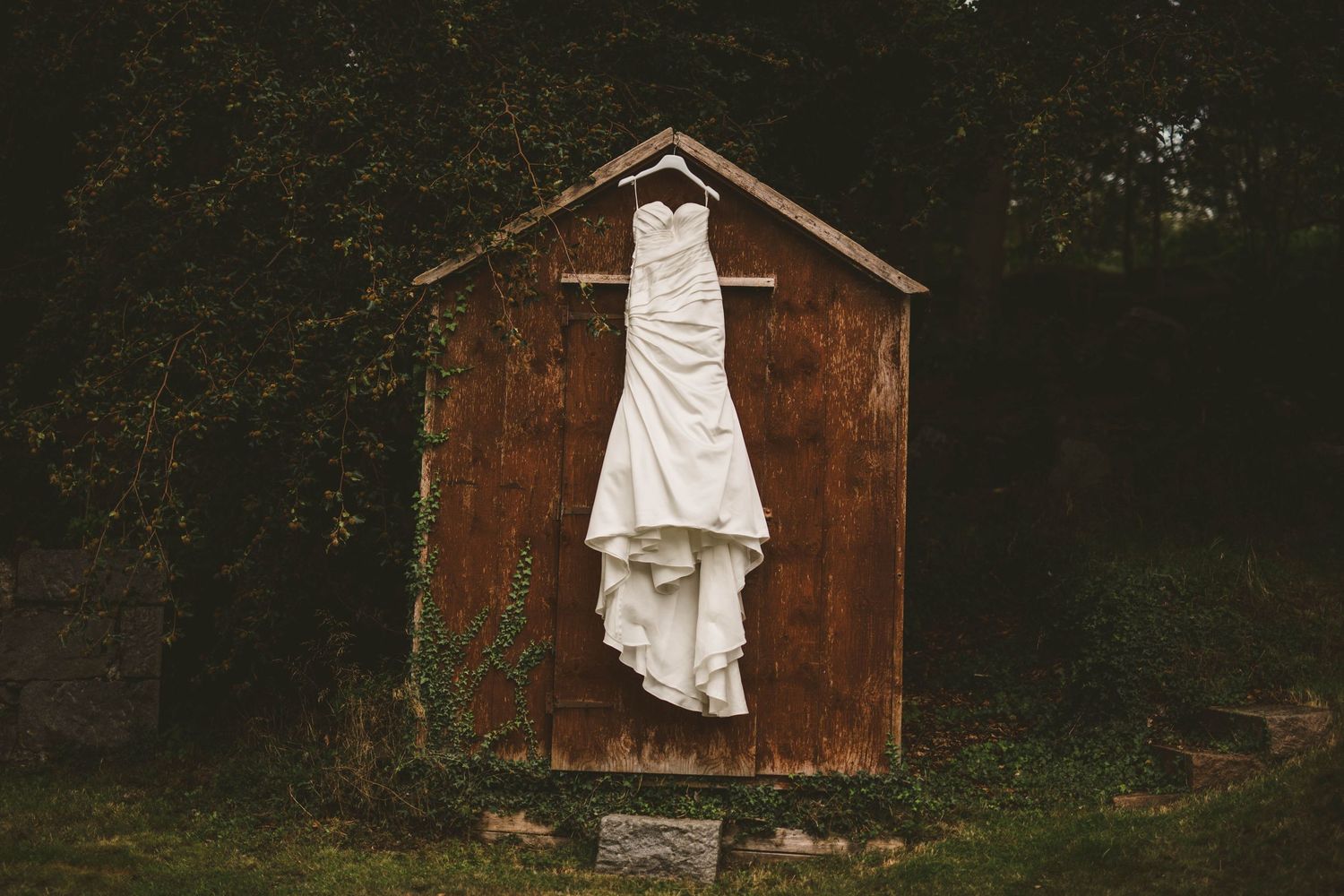 White wedding dress hanging on rustic wooden shed in moody outdoor setting.