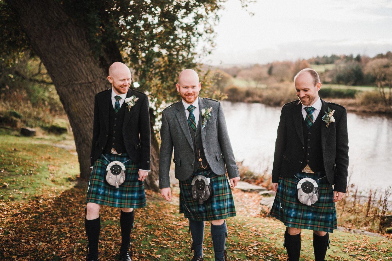 Three groomsmen in matching kilts and formal attire stand together by a lake in an autumn setting.