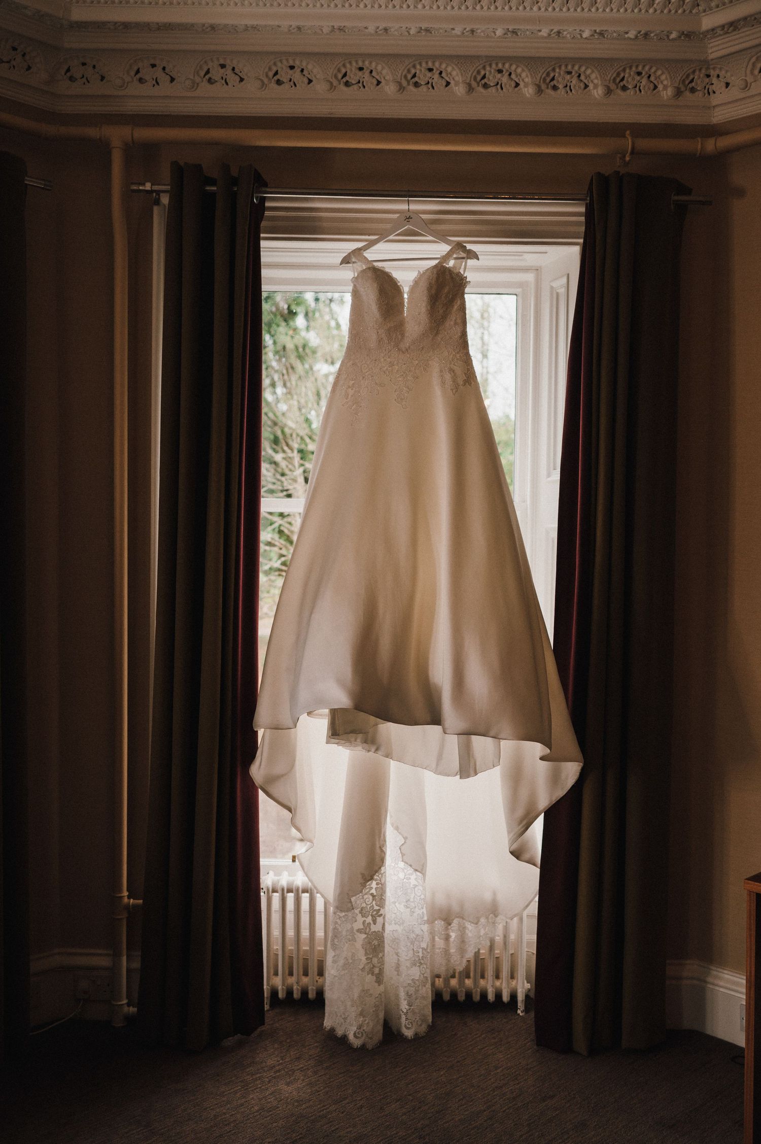 White wedding dress with lace details hanging in front of dark curtains in an elegant room.