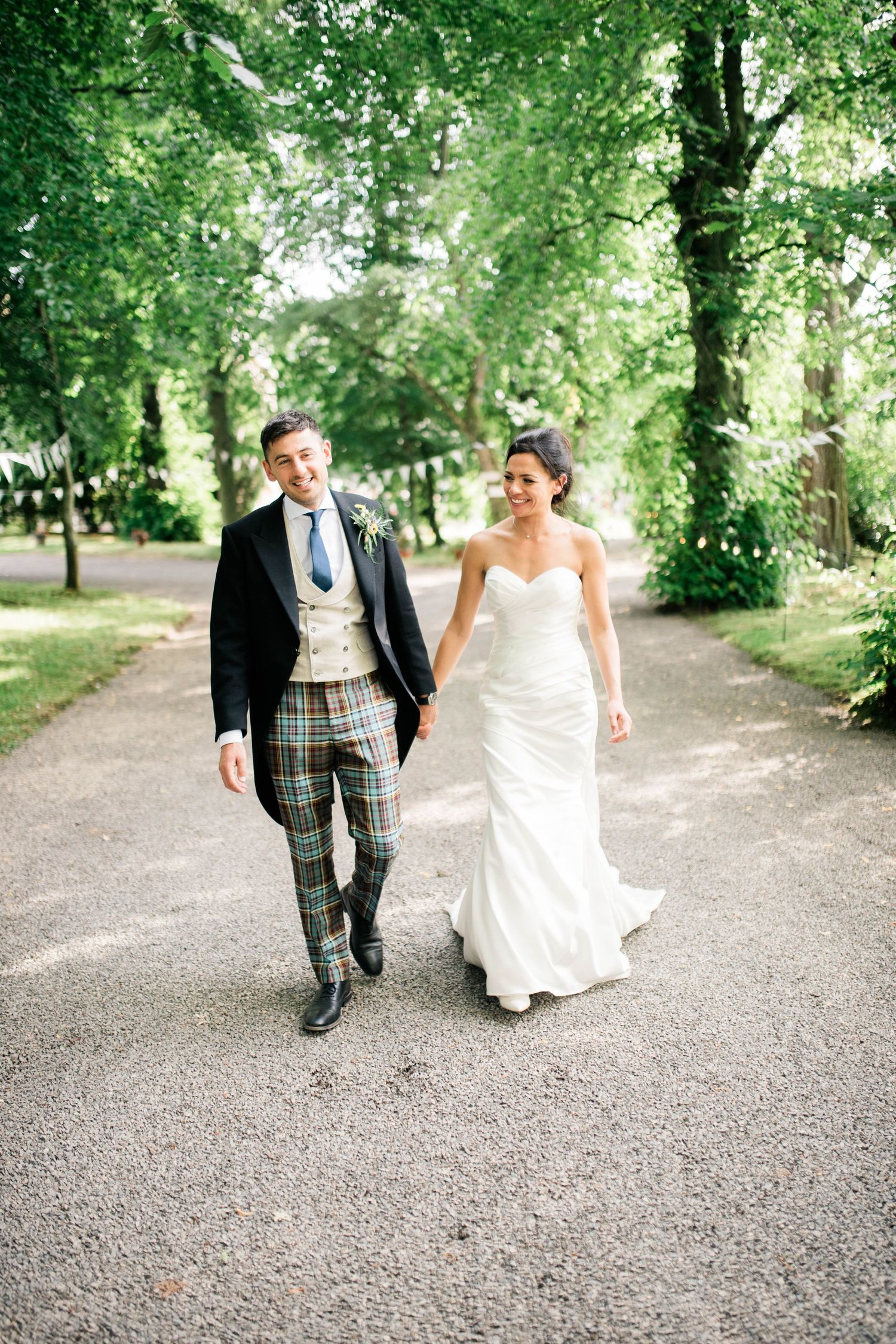 A wedding couple walks down a tree-lined path with the groom wearing traditional Scottish attire.