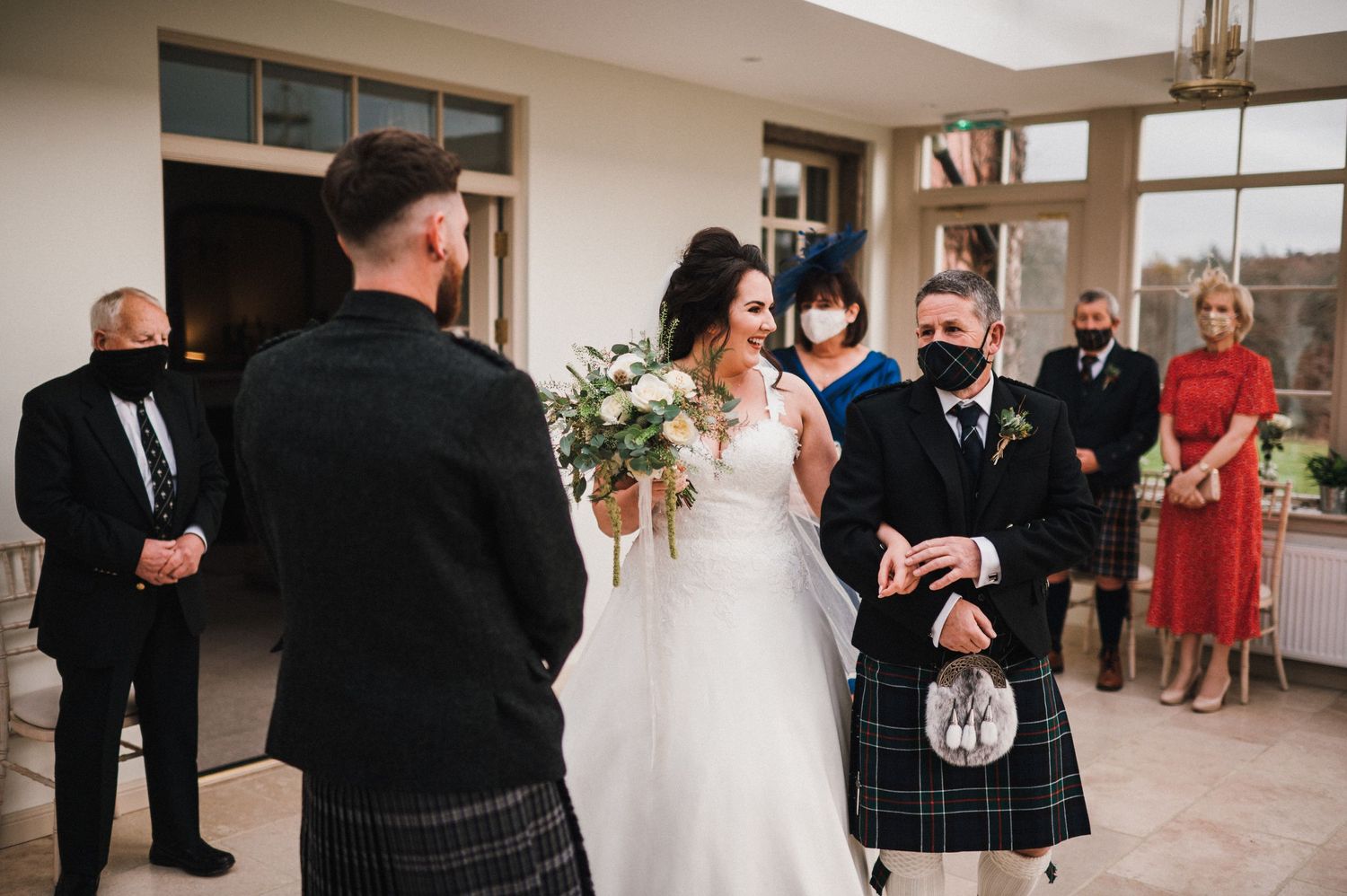 Wedding ceremony in a sunlit room with traditional Scottish formal wear and floral arrangements.