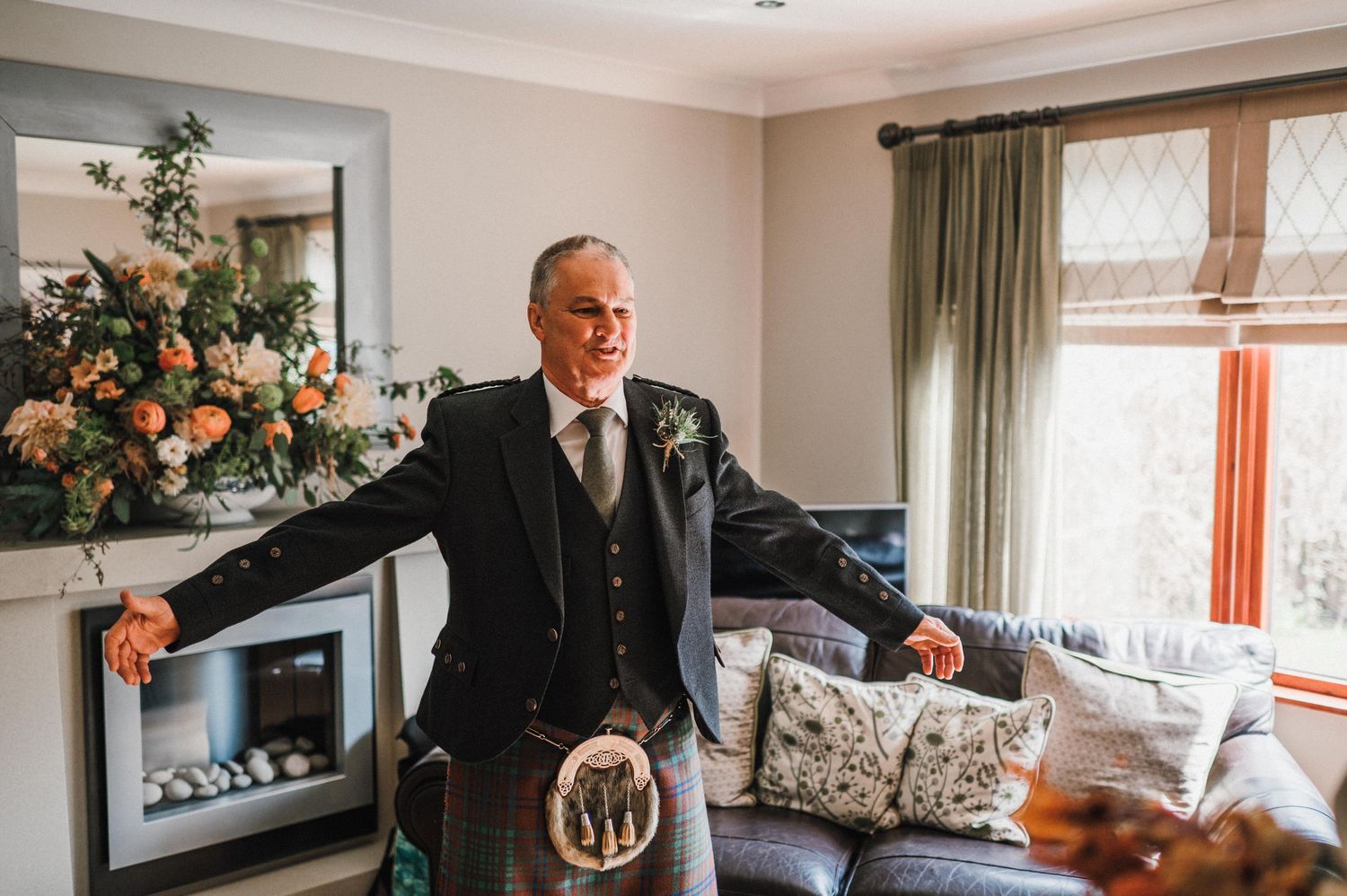 A person in formal Scottish attire poses confidently in a living room with a fireplace and floral decor.