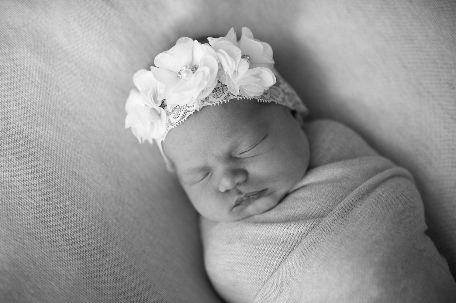 Sleeping newborn baby wearing white floral headband wrapped in soft white blanket.