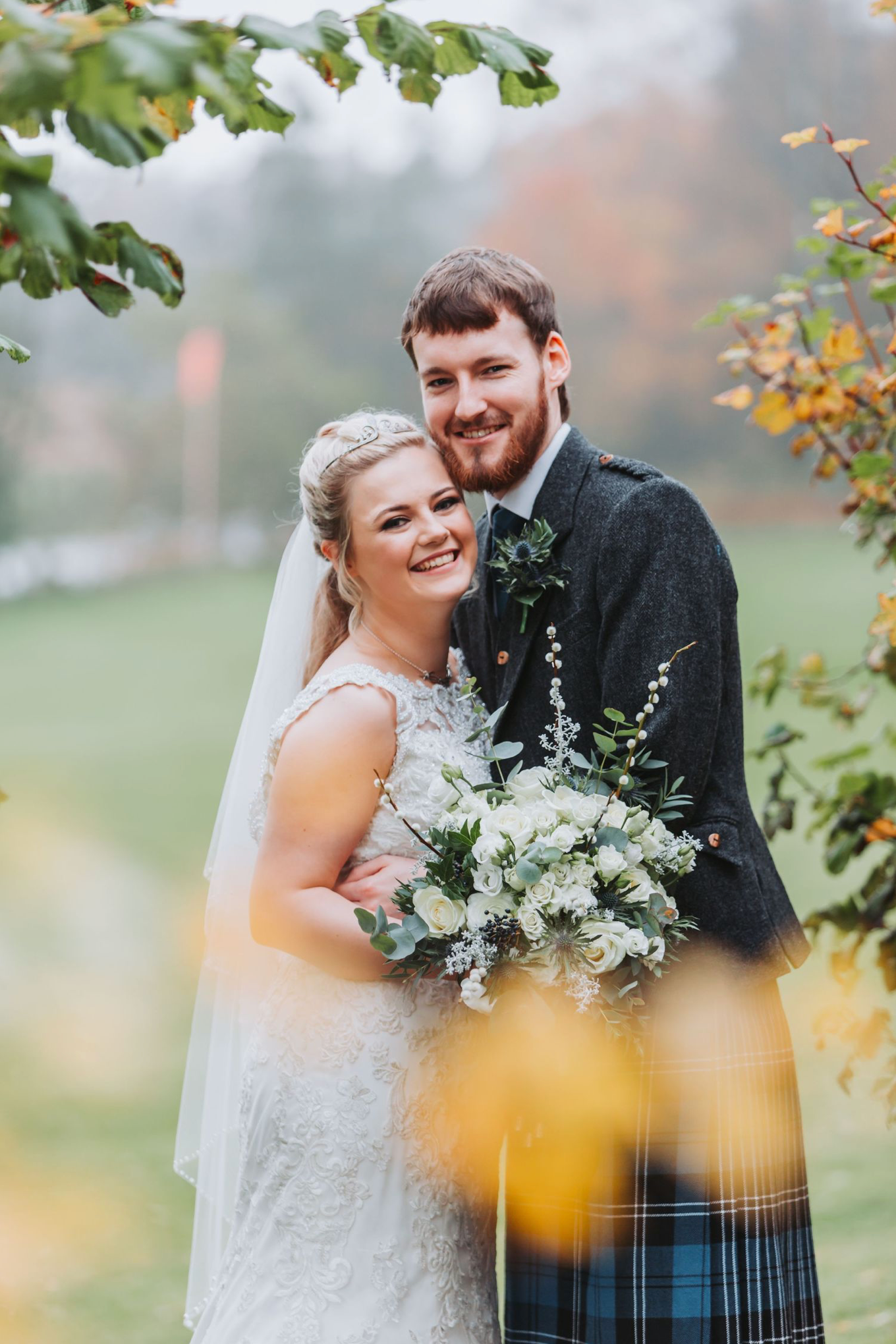 A romantic wedding couple embraces outdoors in autumn, with the bride holding a white floral bouquet against a blurred background.