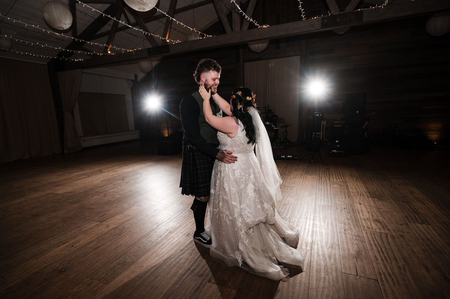A couple dances together on a dark wooden dance floor illuminated by spotlights during their wedding celebration.
