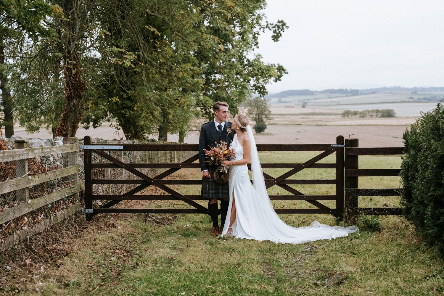 Couple poses by rustic wooden gate with countryside landscape in background.