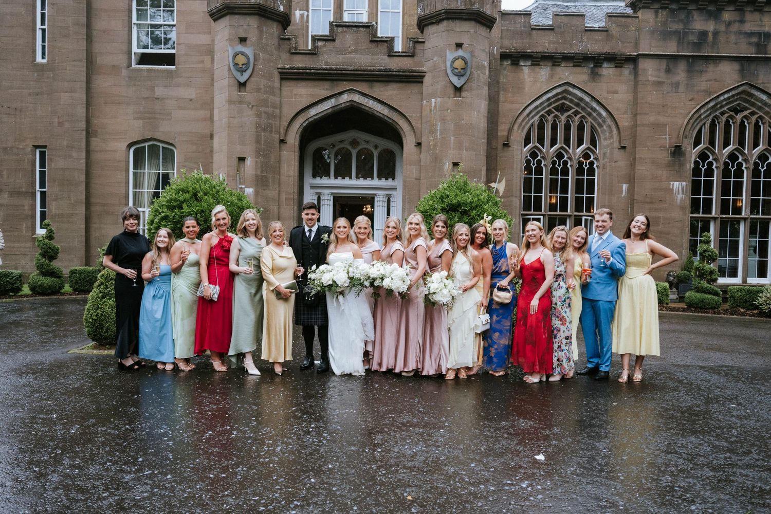 Large wedding party group photo taken outside a historic stone building in the rain.