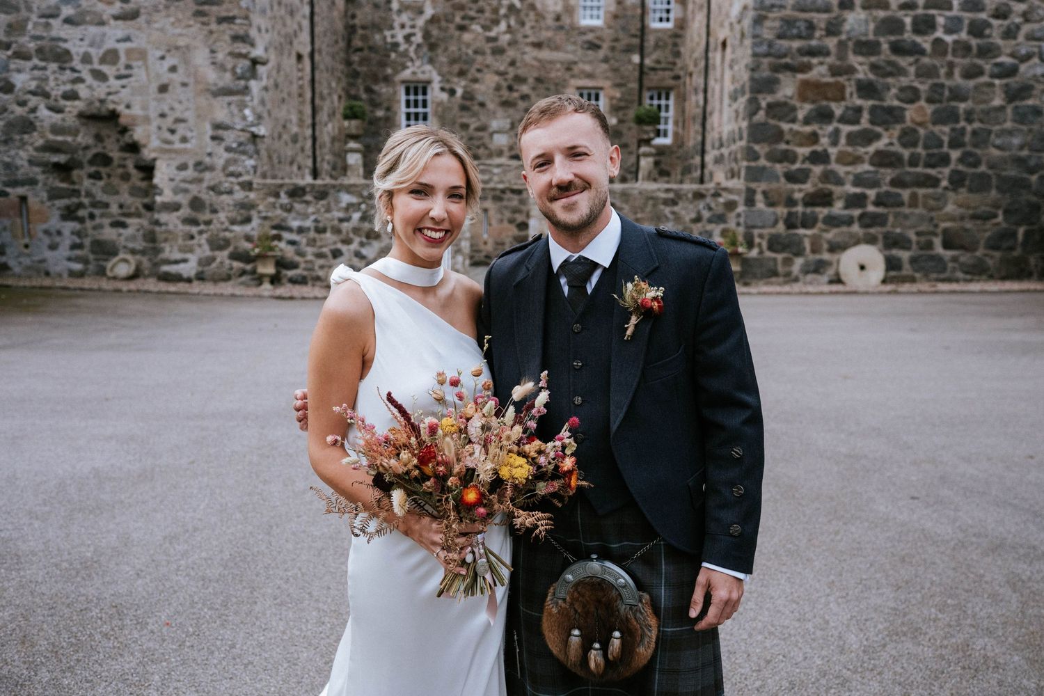 Wedding couple posing in front of a historic stone wall with rustic floral bouquet.