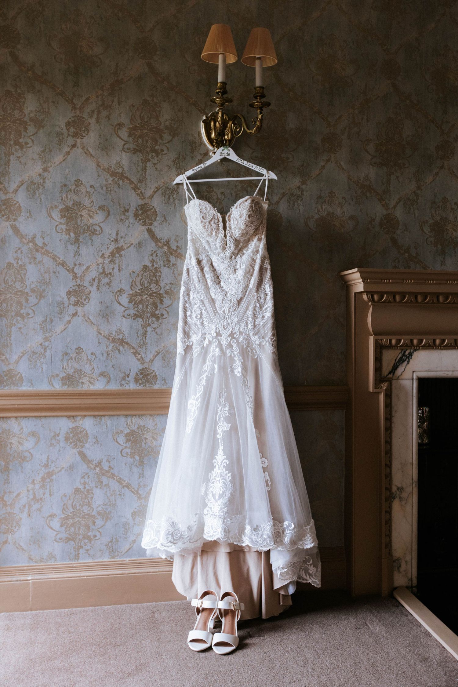 Elegant lace wedding gown hanging against wallpapered wall with vintage sconce lighting.