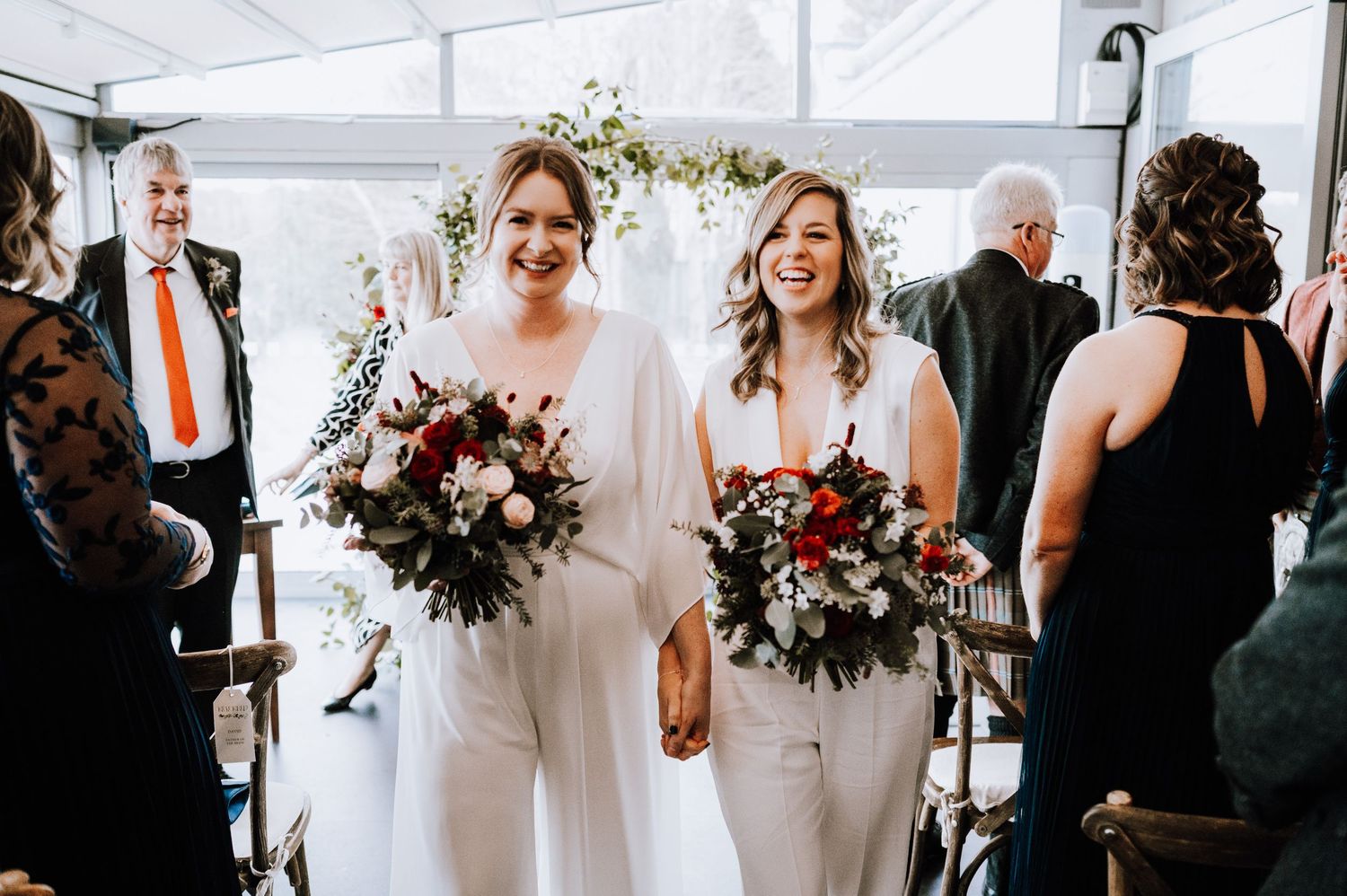 Same Sex Wedding Photography. Two people in elegant white outfits hold beautiful burgundy bouquets under a white tent.
