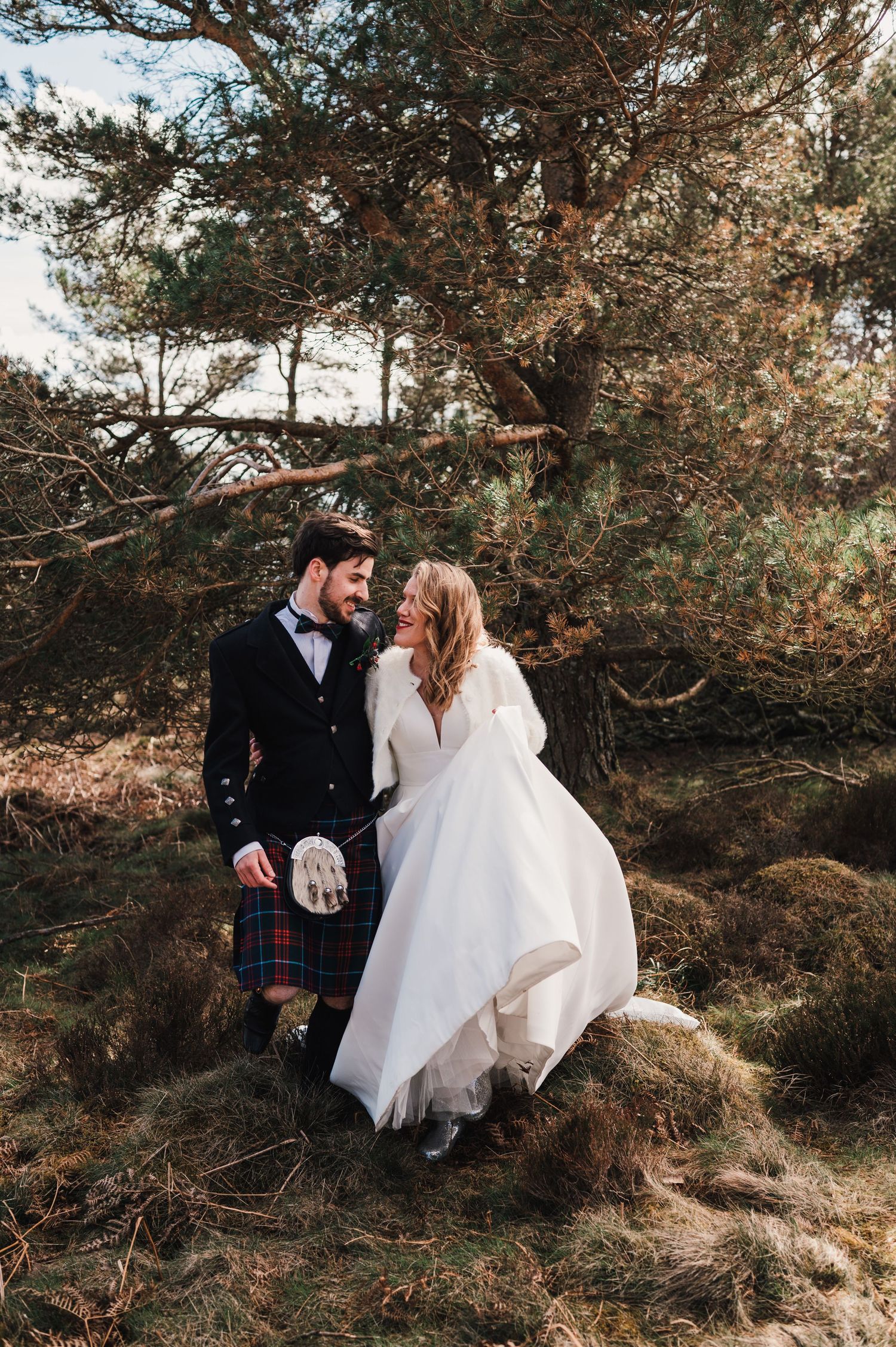 A couple in traditional Scottish wedding attire embraces among pine trees on a rustic hillside.