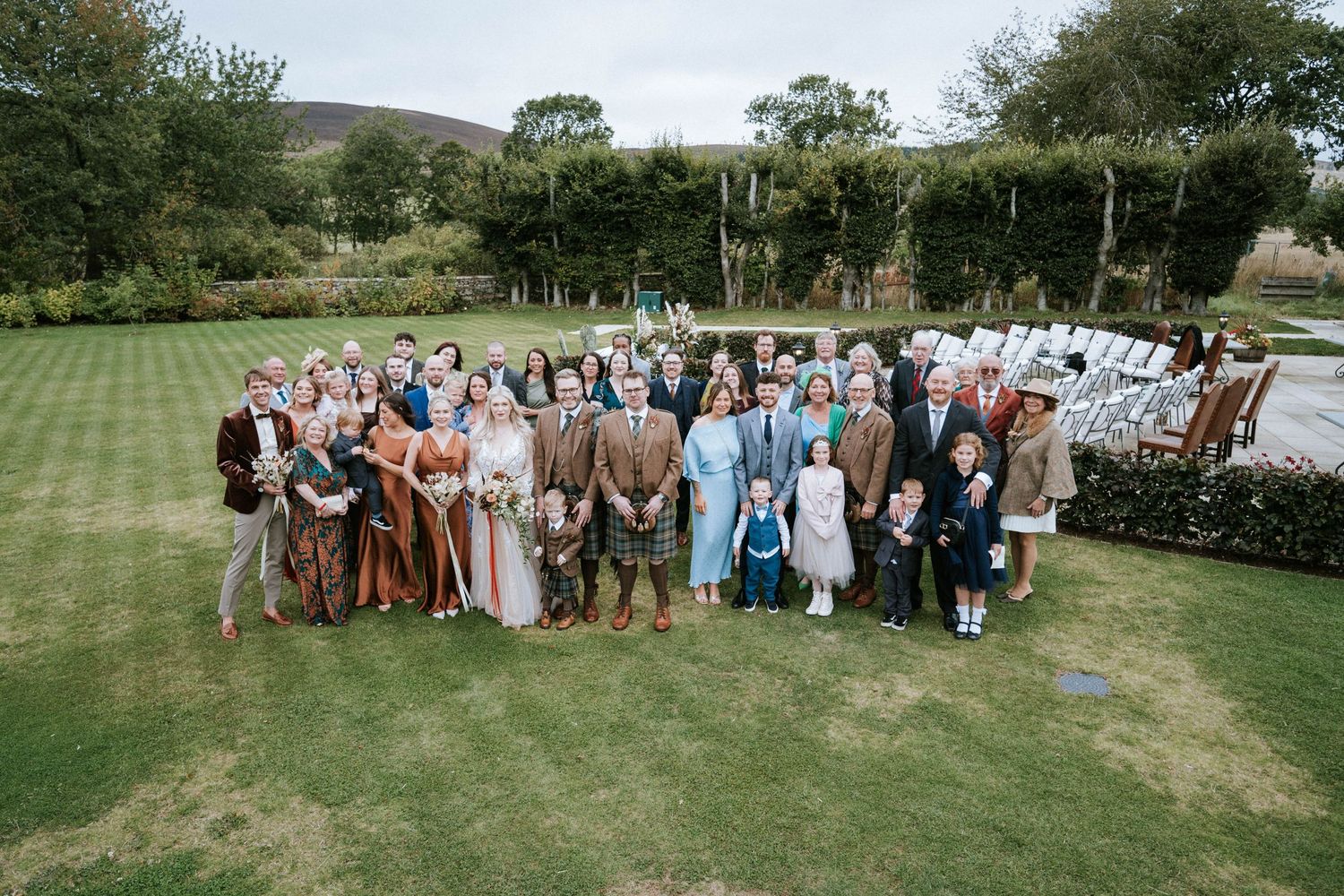 Large wedding party group photo on a manicured lawn with guests wearing mix of formal attire and traditional kilts.
