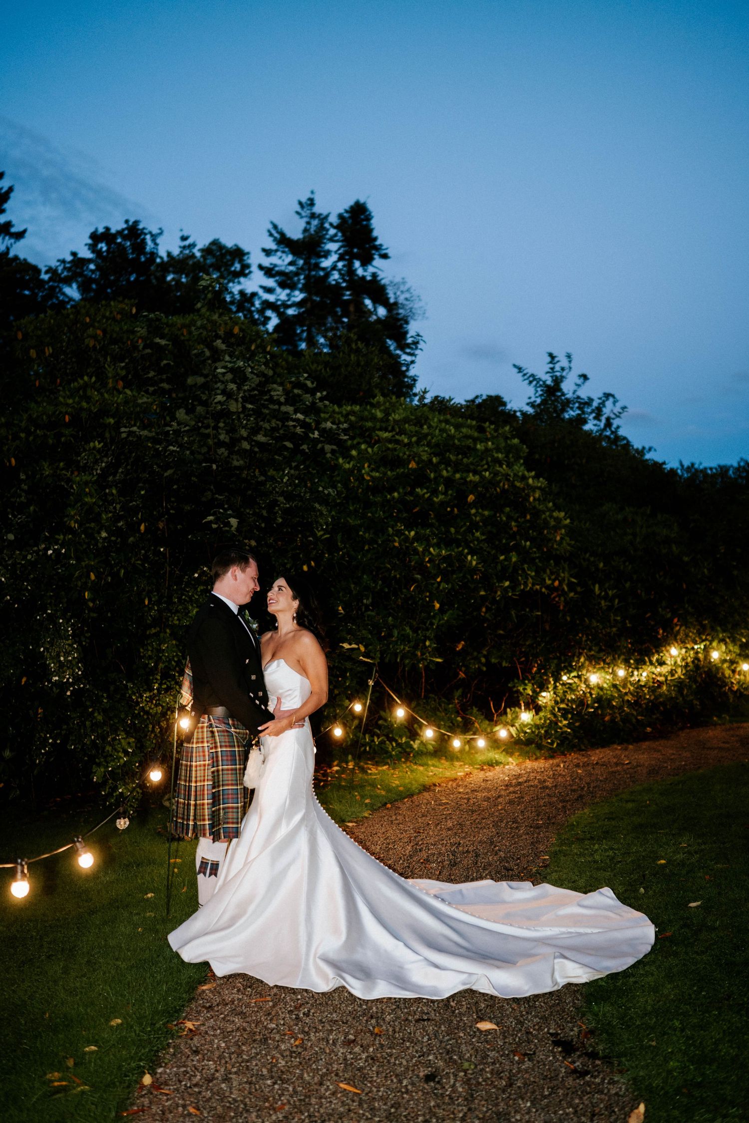 A romantic wedding photo of a couple in Scottish attire beneath string lights and trees at dusk.