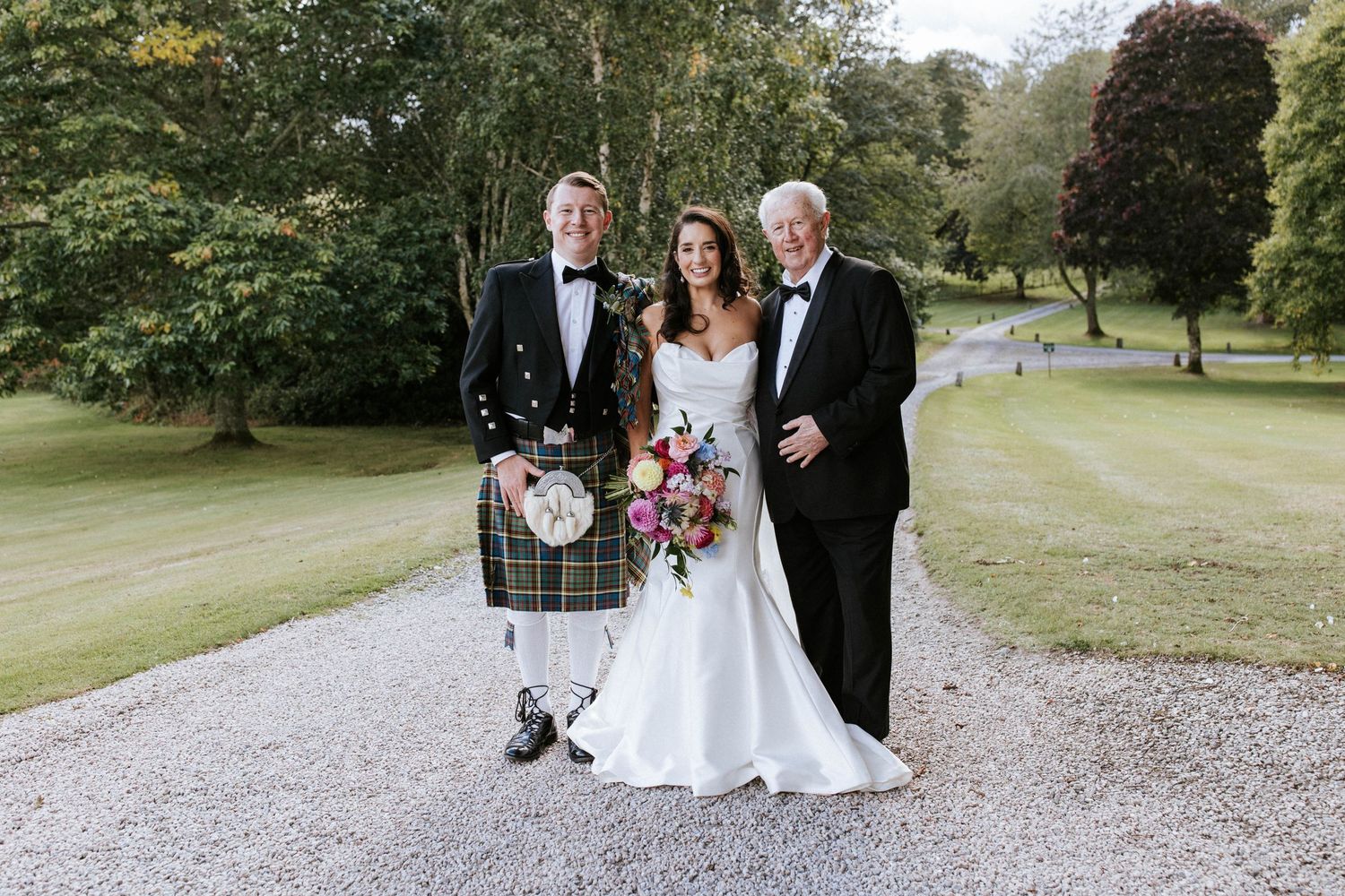Three formal wedding attendees pose on gravel path with one wearing traditional Scottish kilt.