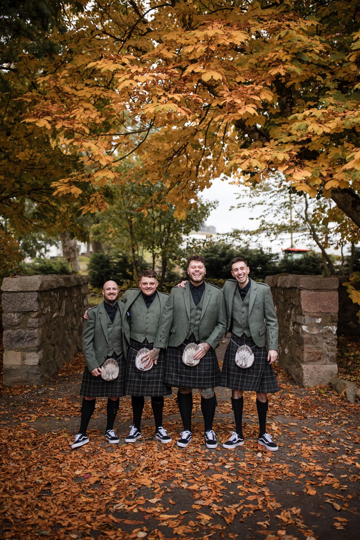 Group of groomsmen sitting on stone wall with autumn leaves and trees in background wearing matching gray suits.