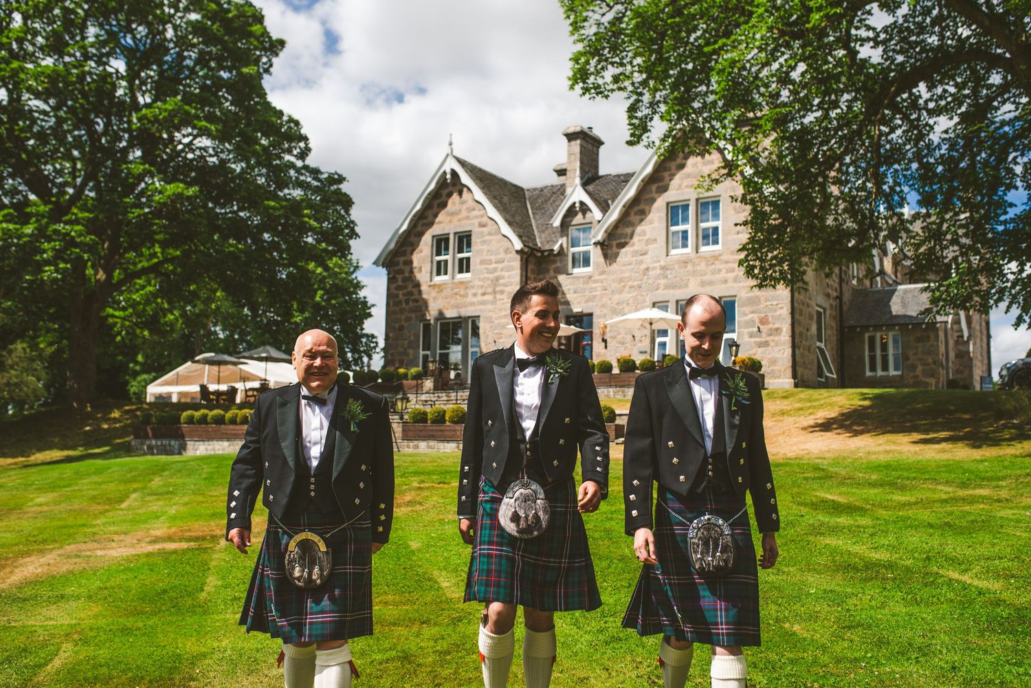 Groomsmen in traditional Scottish kilts pose on lush green lawn with stone building in background.