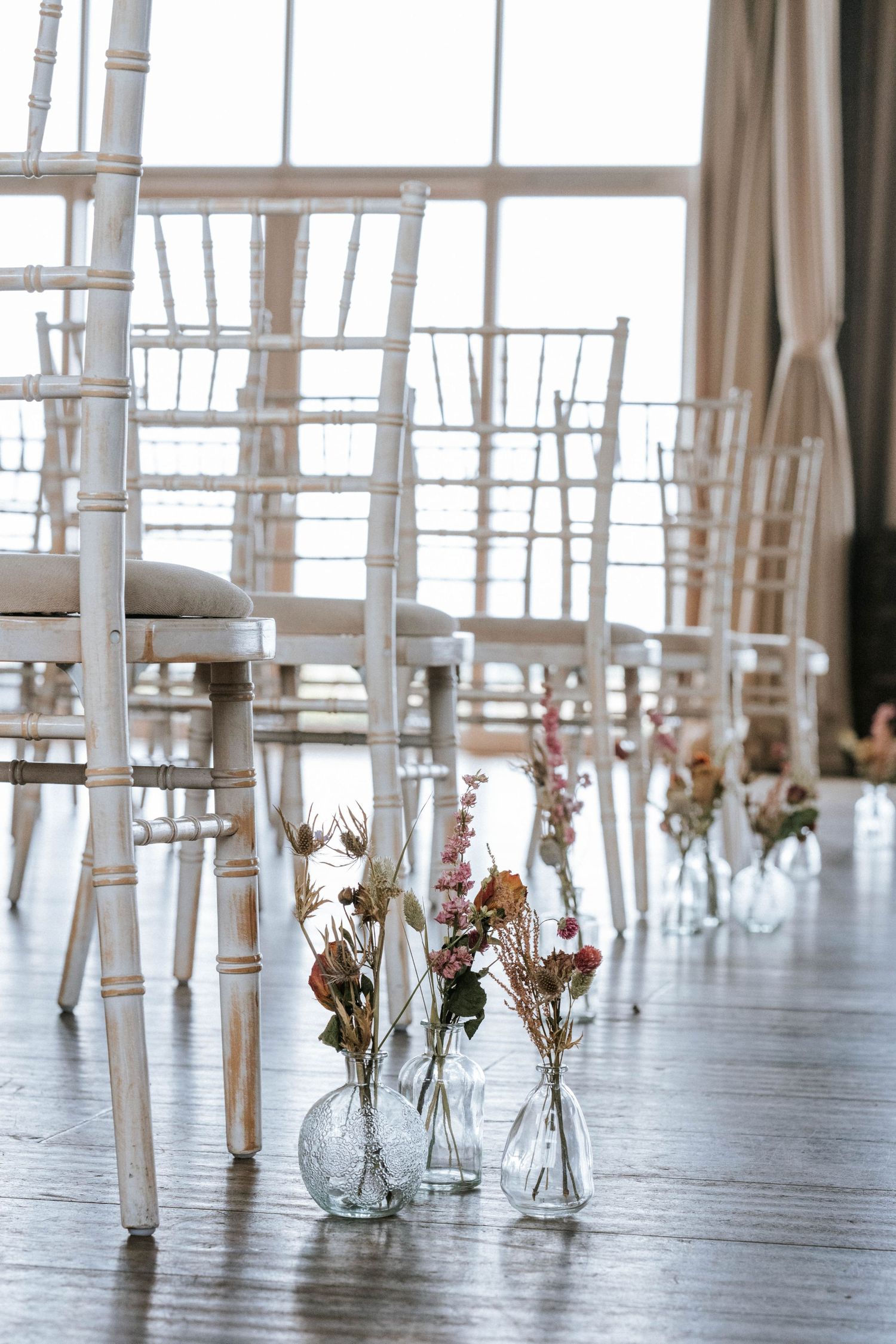 Long rows of delicate flower vases with small blooms line a polished wooden floor in an airy event space.