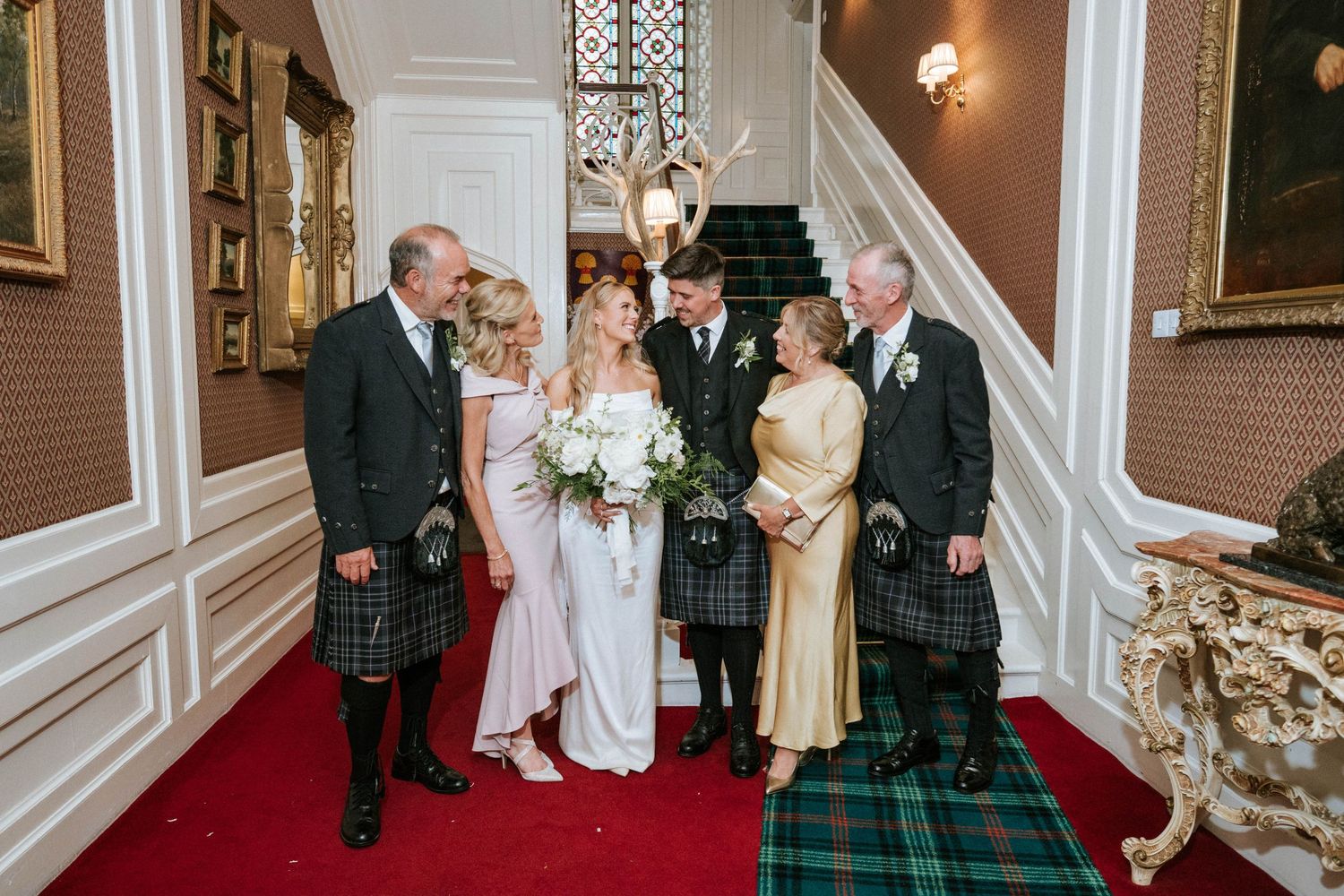 Wedding party in traditional Scottish attire poses on red-carpeted stairs in an ornate venue.