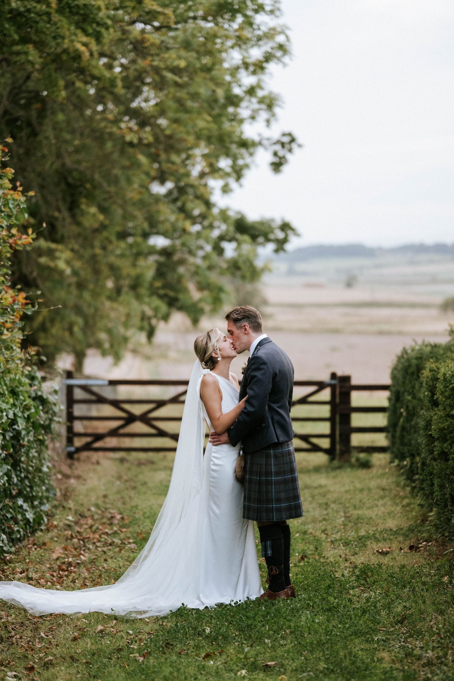 A romantic wedding couple shares intimate moments in a scenic countryside setting with rustic wooden fences and green foliage.