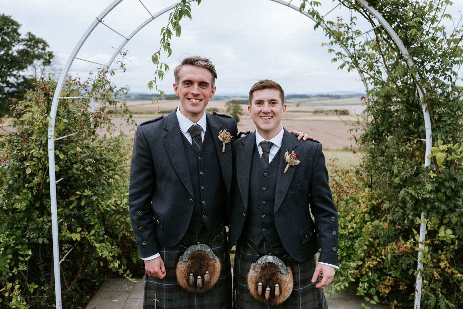 Two men in formal suits and kilts stand under a garden arch with scenic countryside views.