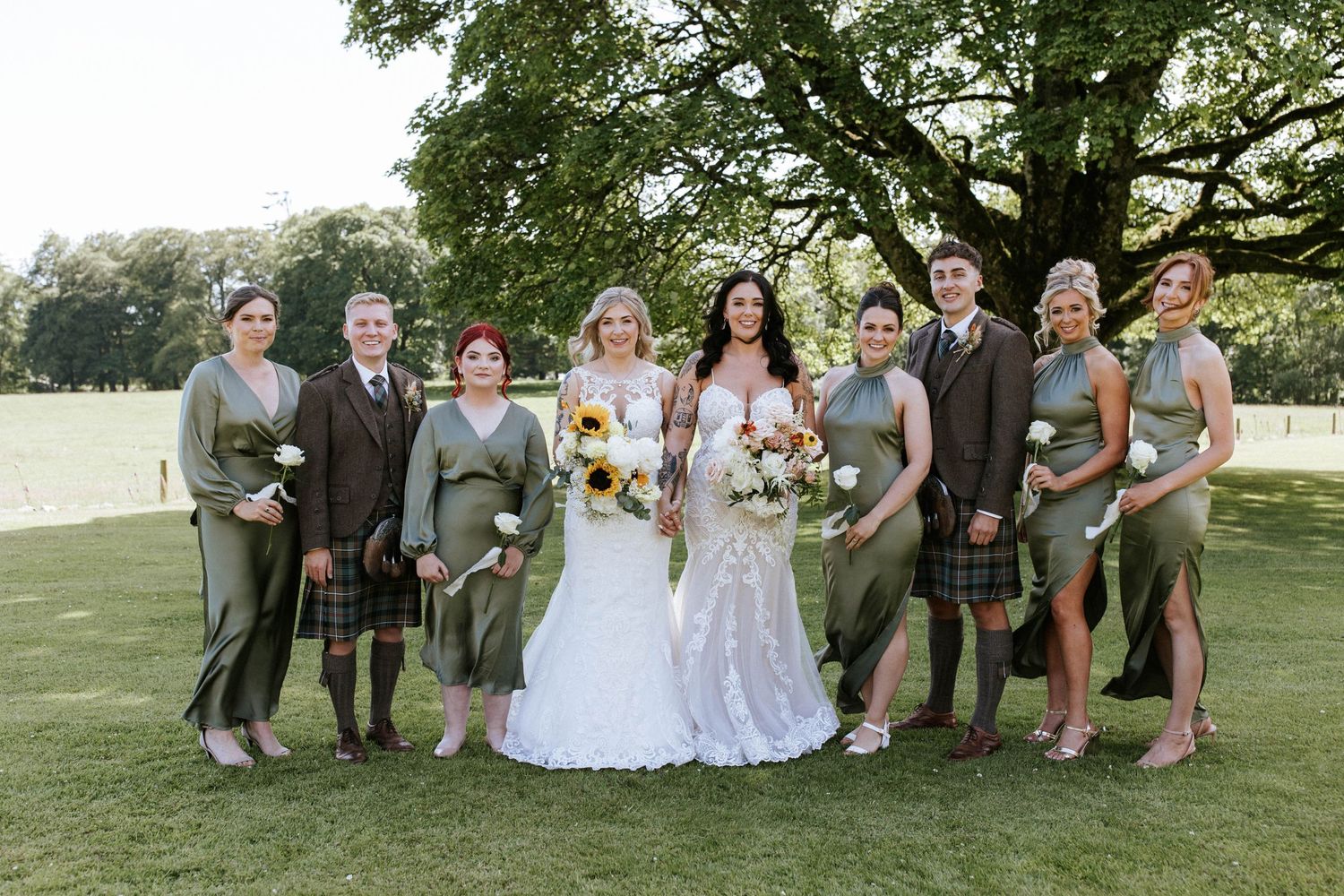 Wedding party members in mix of formal wear and kilts pose together on grassy hillside.
