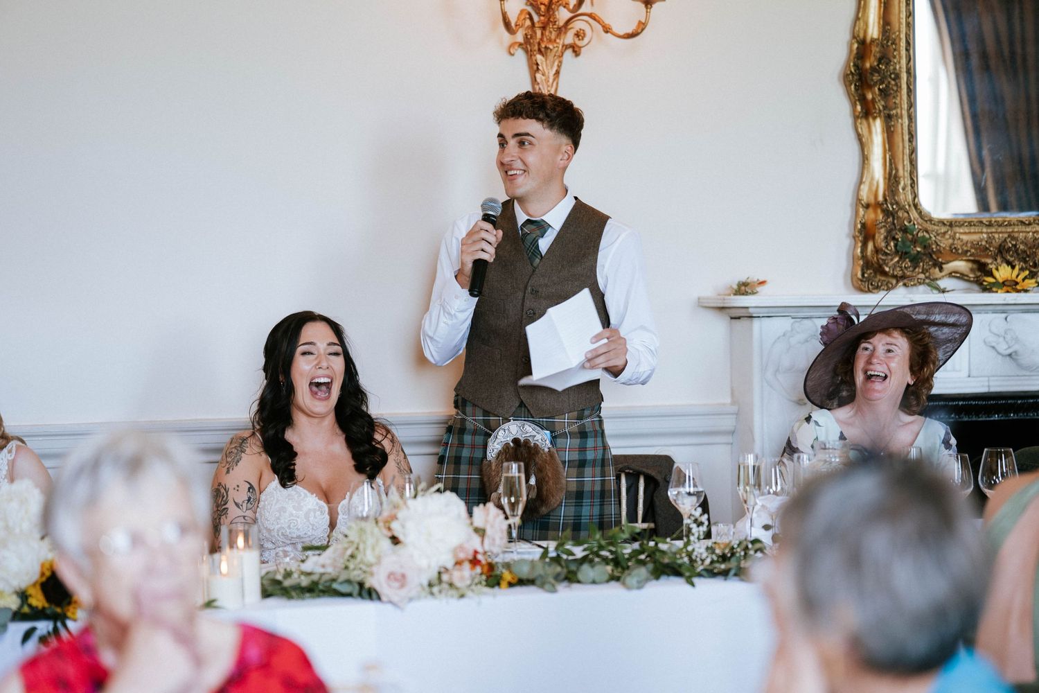 Joyful laughter erupts during wedding reception speeches at an elegant venue with chandelier.