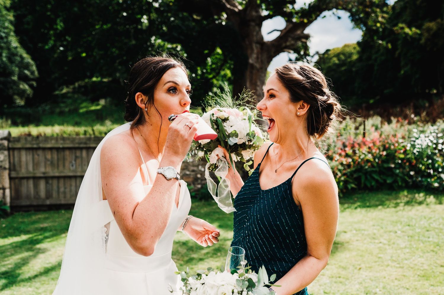 Two people in black and white formal attire share a candid laughing moment together in a sunny outdoor garden setting.