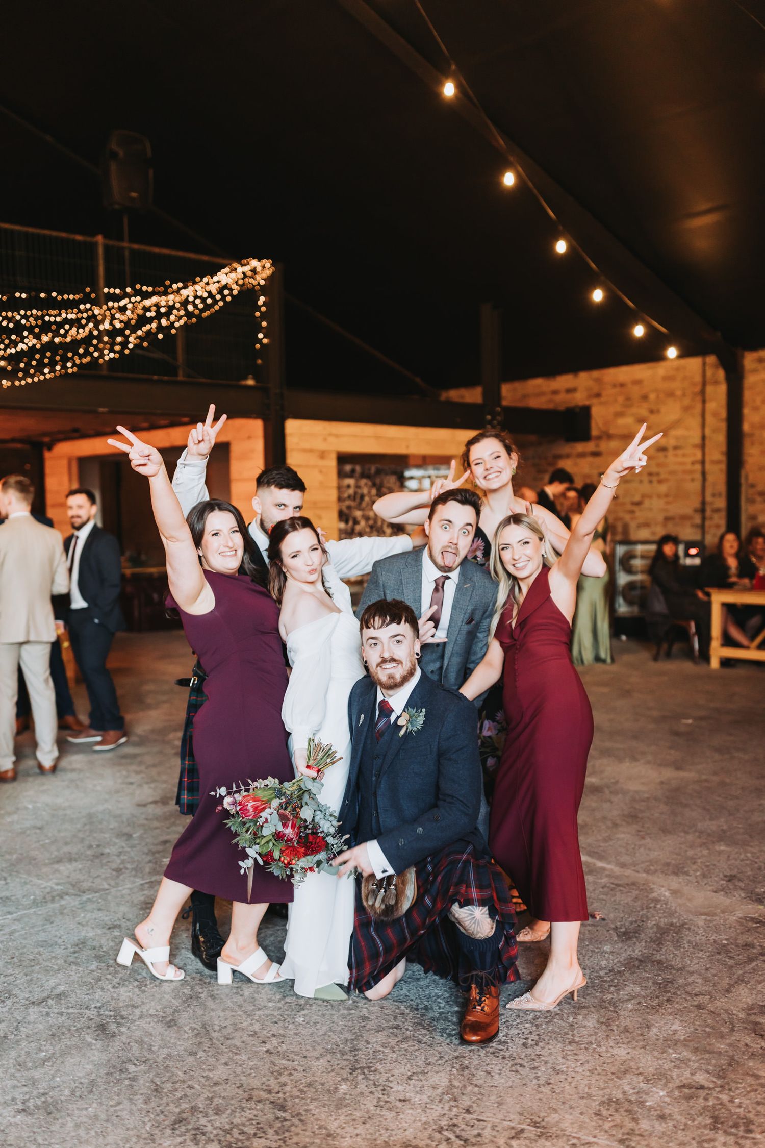 Wedding party members pose playfully on the ground under string lights at an outdoor evening reception.