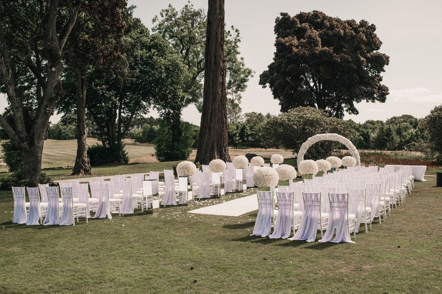 Wedding ceremony setup with white chairs and floral arch in a garden setting.