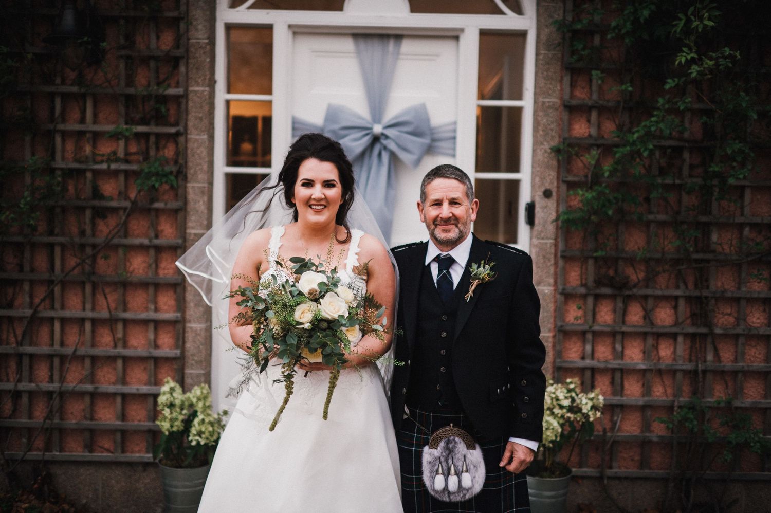 A wedding couple poses in front of a brick building with climbing vines and white bow decor.