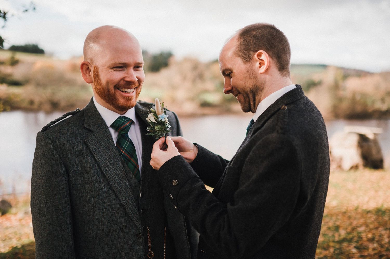 Two people in formal attire adjusting boutonnieres by a lake.