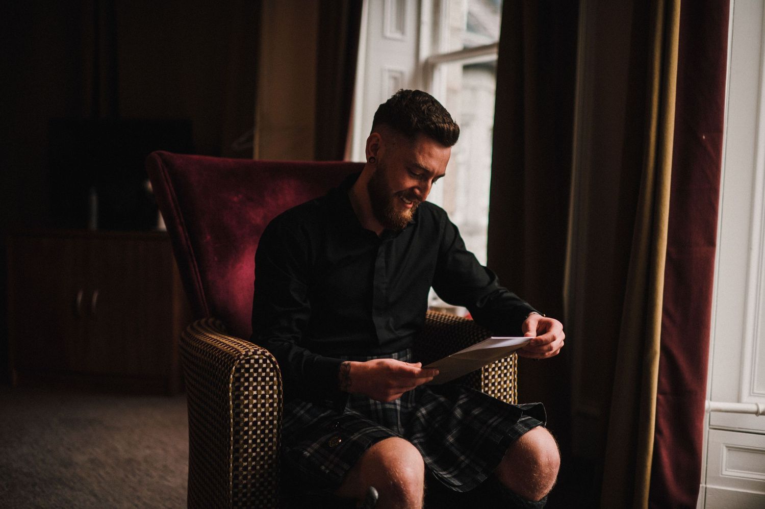 Person reading letters while seated in a burgundy velvet chair near a window.